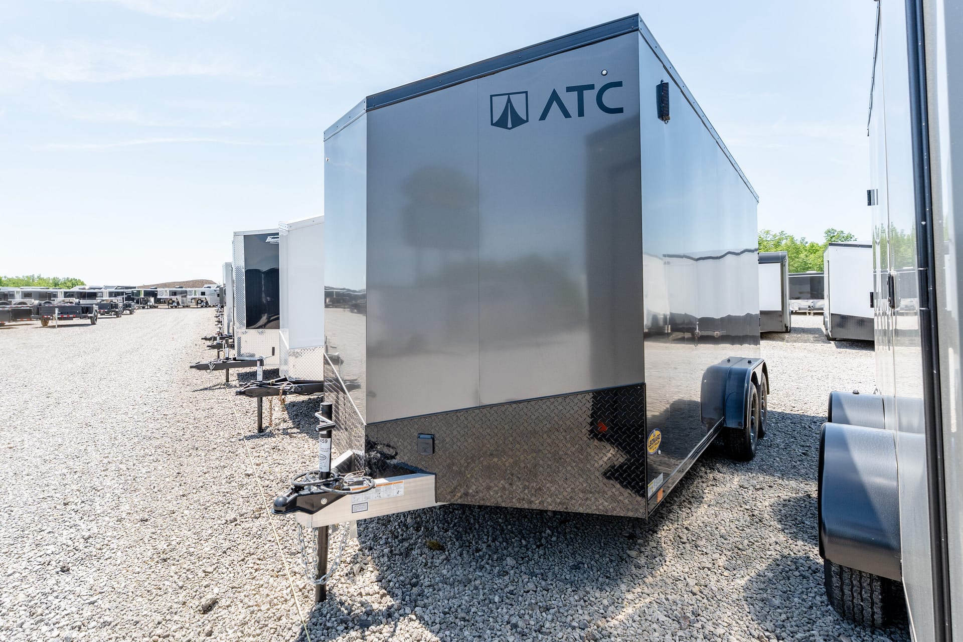 Row of enclosed cargo trailers on a gravel lot, foreground shows a tall gray ATC trailer with reflections on its shiny panels under a clear blue sky.