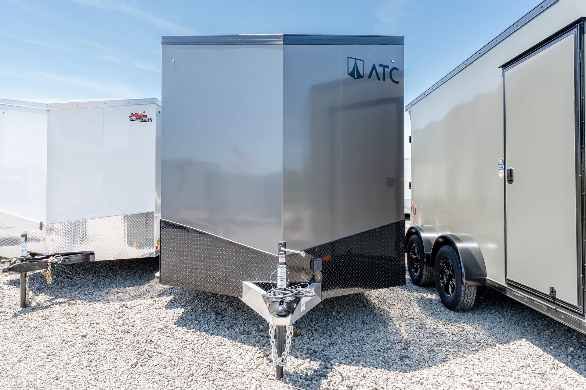 Row of large enclosed cargo trailers on a gravel lot under a clear blue sky; center gray ATC trailer stands out with logo visible.