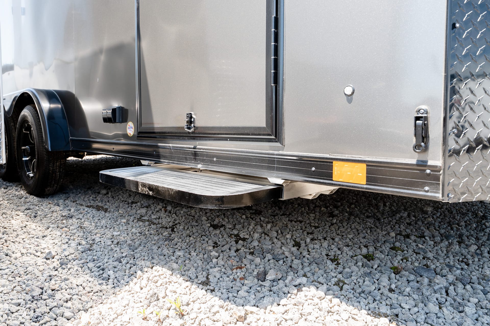 Side view of a silver enclosed trailer with a small access door, fold-down step, and wheel on gravel ground.