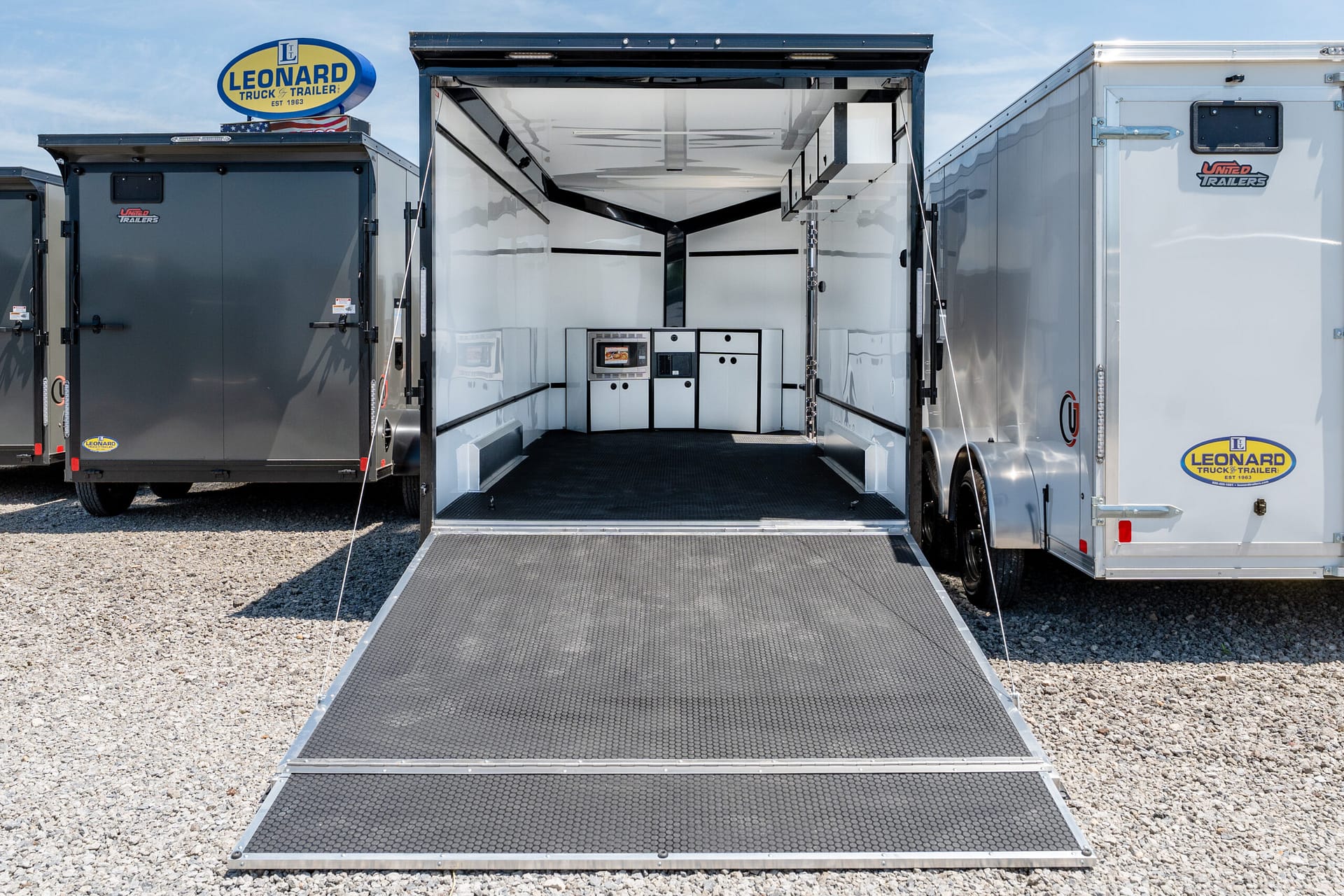 Open trailer with a metal ramp extended to a gravel lot; white interior with cabinets and a built-in kitchenette visible inside. Leonard Truck & Trailer sign on the left.