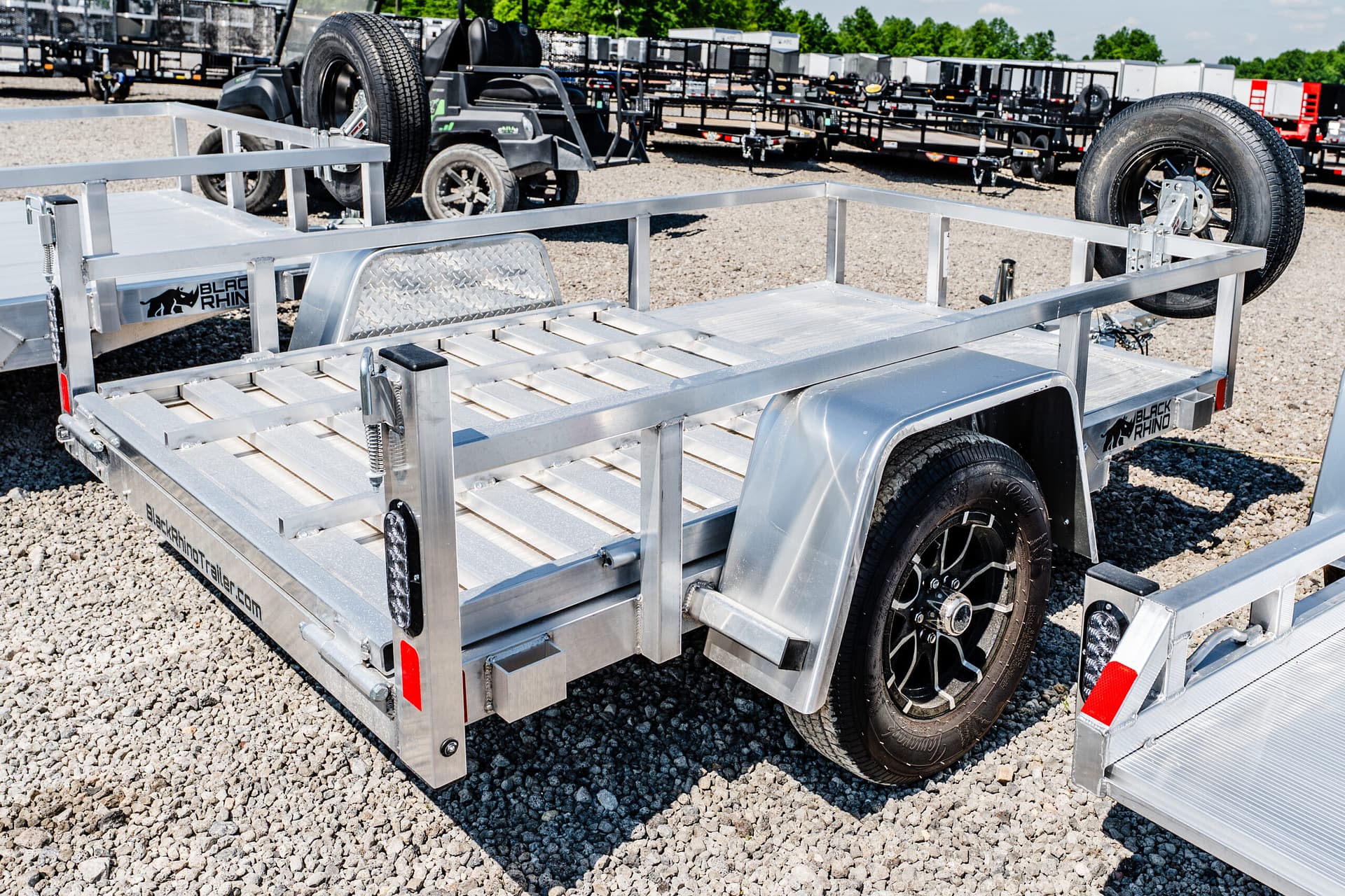 Aluminum utility trailer with a mounted spare tire on the side, parked on a gravel lot with others in the background (Black Rhino branding).