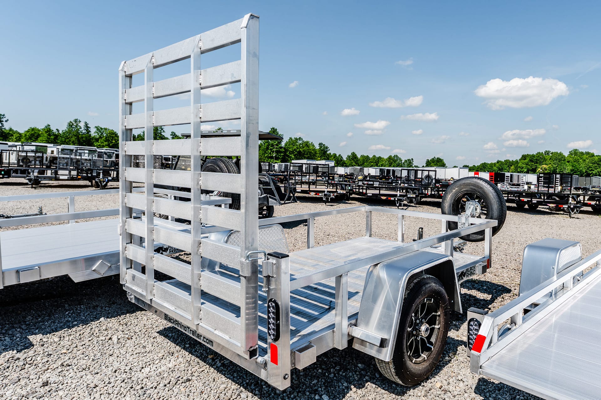 Silver aluminum utility trailer with a ramp gate in a gravel lot, other trailers lined up in the background under a clear blue sky.