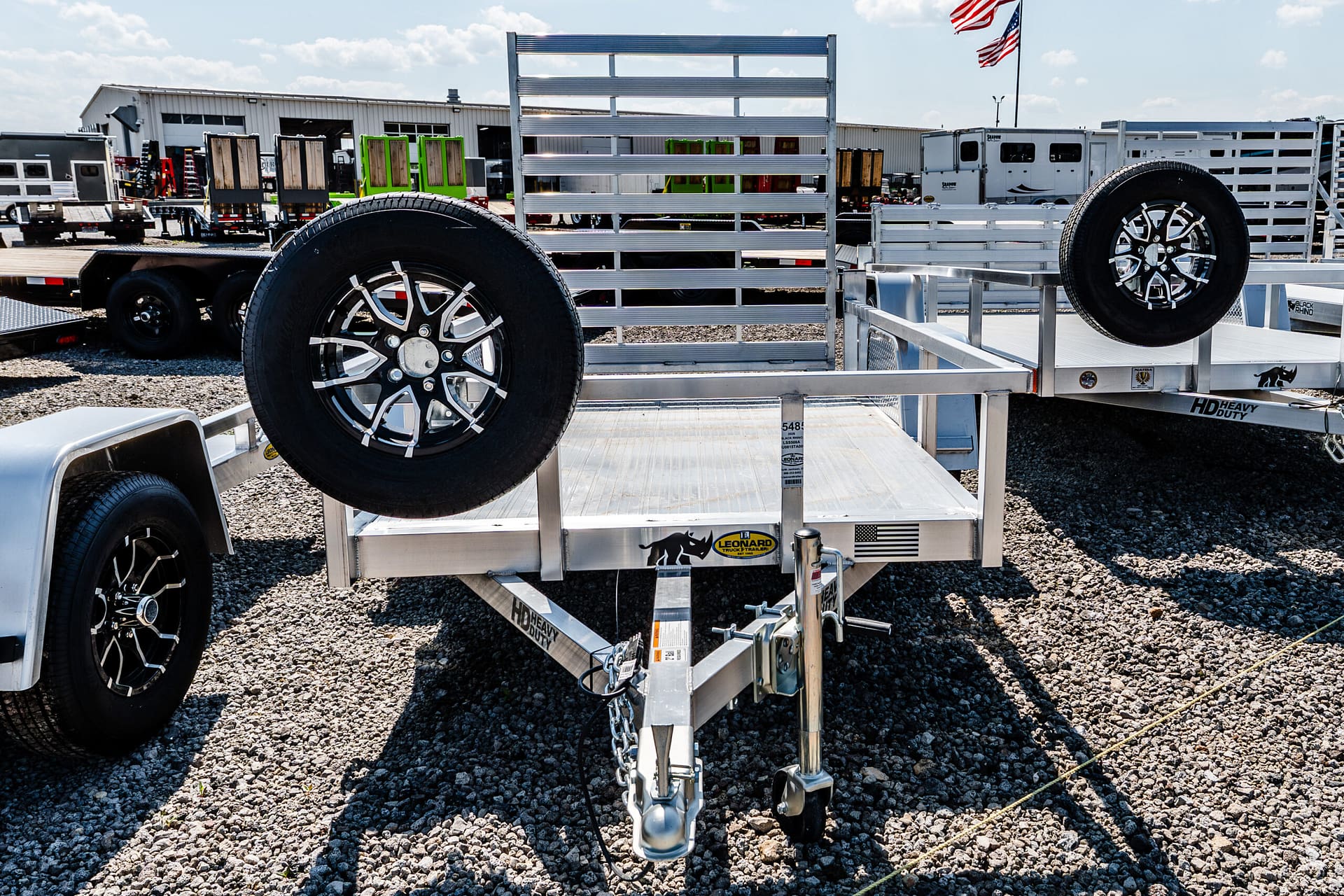 Close-up of a silver heavy-duty trailer with a mounted spare tire and aluminum ramp, parked on a gravel lot at a trailer dealership.