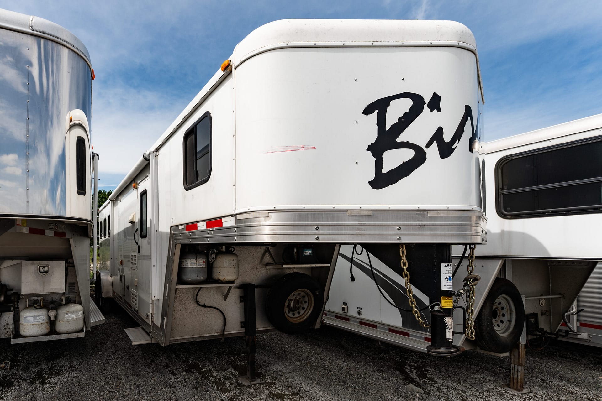 White semi-trailers parked closely together on gravel under a blue sky, with a large black one-letter graphic on the side of the central trailer.