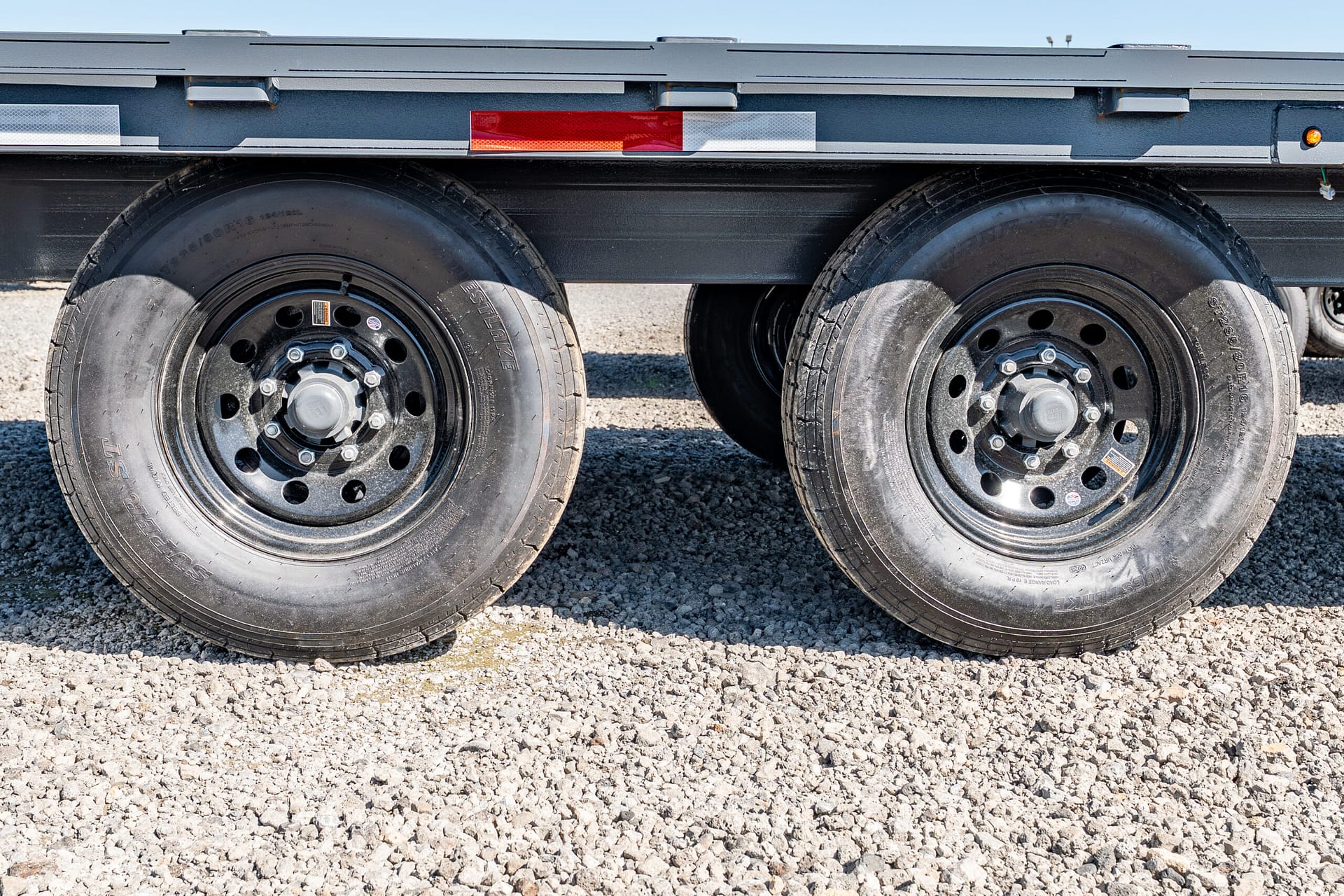 Dual trailer wheels and metal hubs on a gravel surface with red/white reflective tape along the trailer frame, outdoor setting
