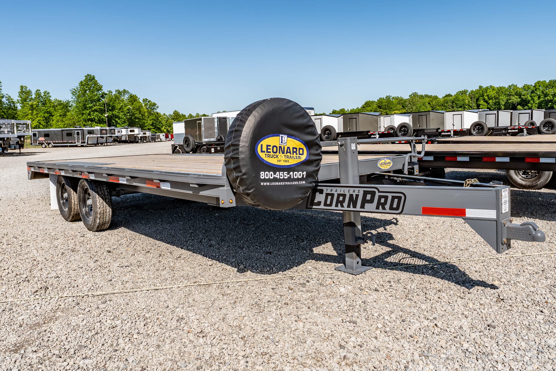 Front view of a large flatbed trailer with a spare tire cover displaying the Leonard Trucks & Trailers logo in a gravel lot, with numerous trailers and greenery in the background under a clear blue sky.