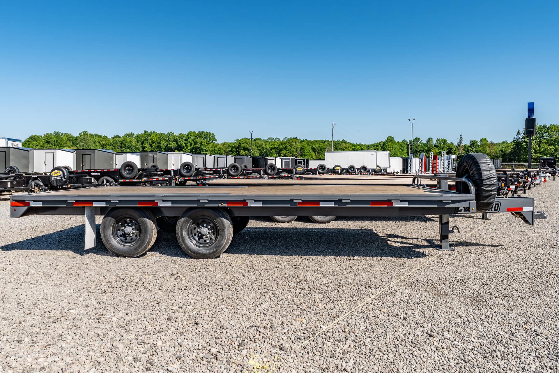 Row of flatbed trailers with spare tires parked in a gravel lot under a clear blue sky, with trees in the background.