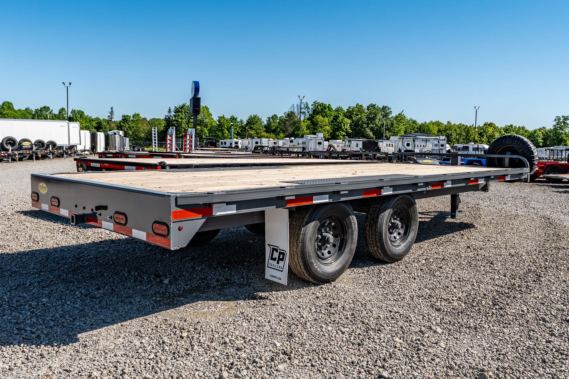 Wide view of a trailer yard with flatbed trailers and trucks under a clear blue sky, Gravel lot and rows of equipment visible in distance