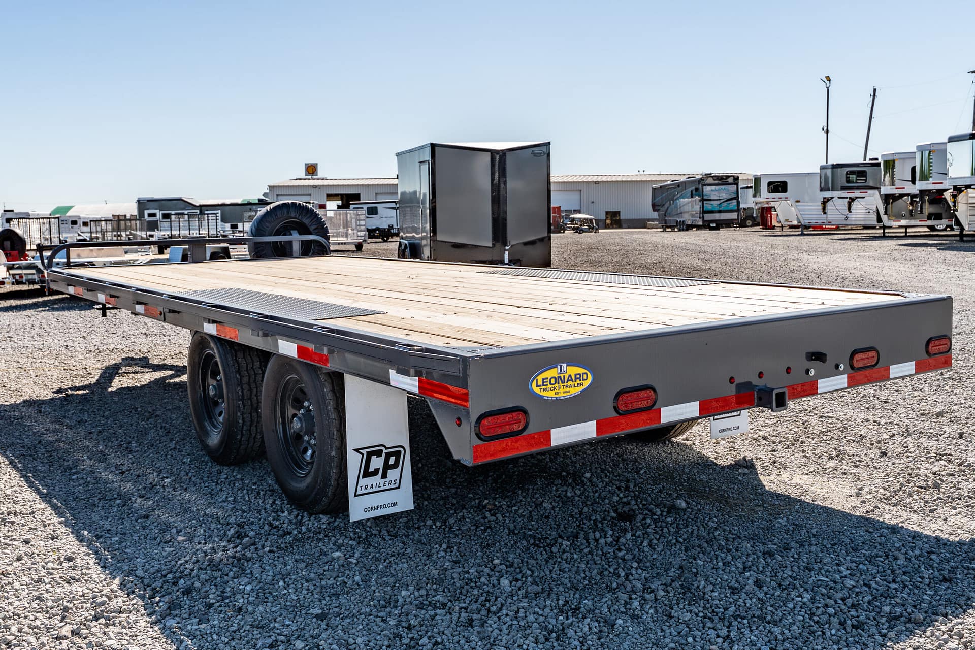 Flatbed trailer in a gravel lot with other trailers and a warehouse in the background.