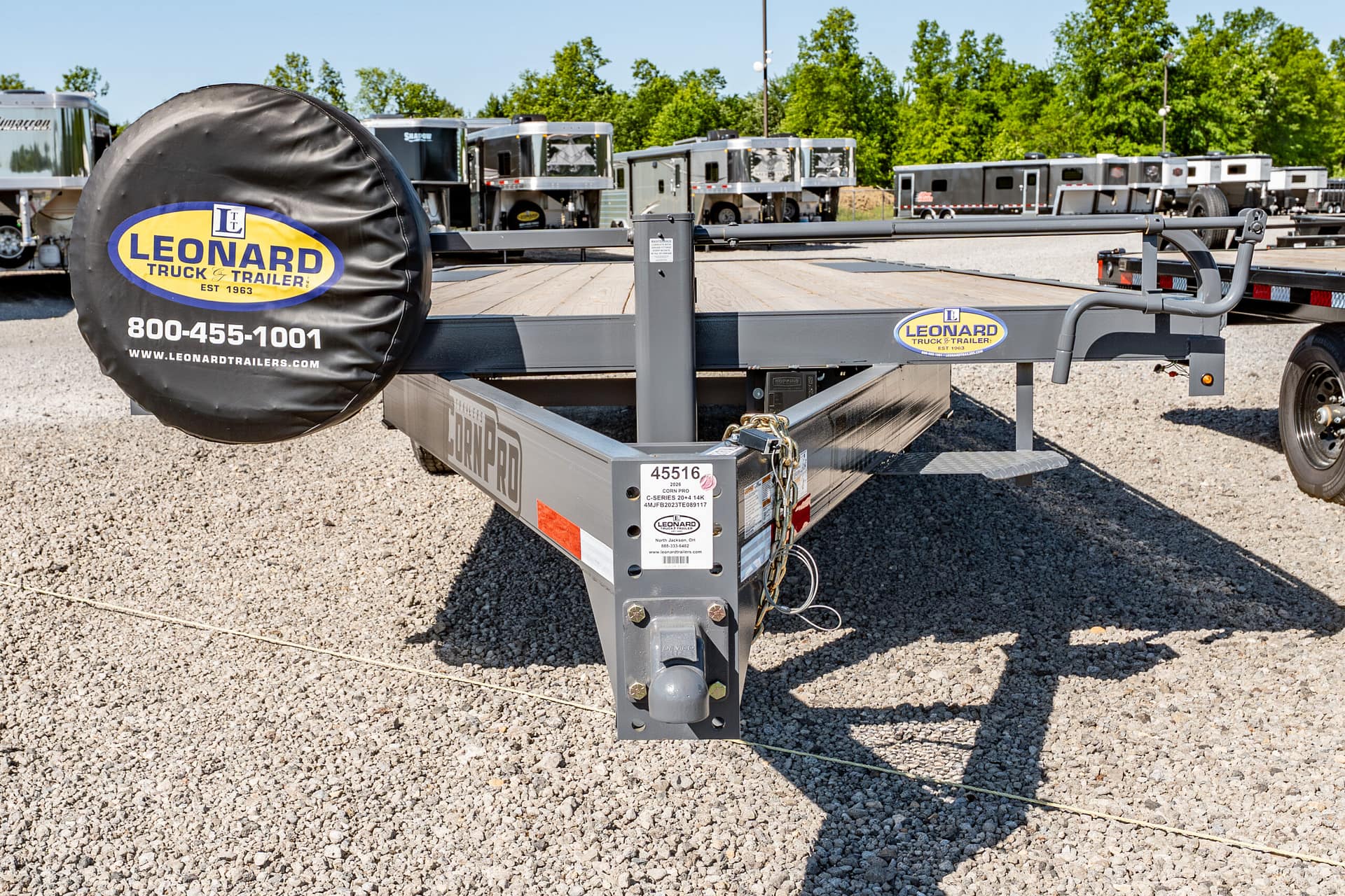 Front section of a Leonard trailer frame with a large spare tire cover bearing the Leonard logo and contact number, on a gravel lot with other trailers in the background.