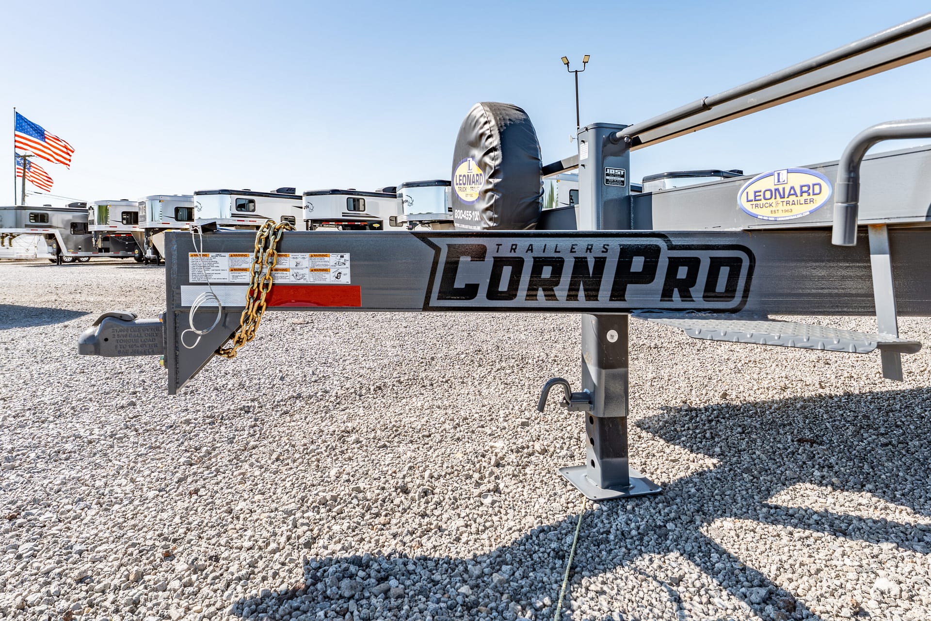 Front section of a CornPro trailer frame with a spare tire cover, chain, and Leonard Trailer logo on a gravel lot, American flags in the background.