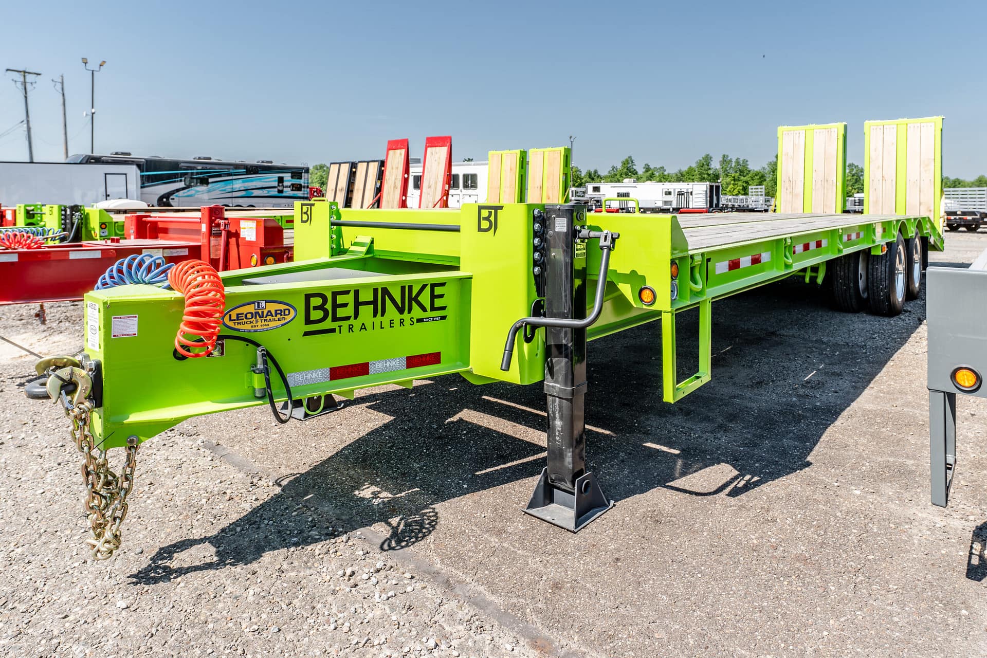 Bright lime-green Behnke trailer with a wooden deck and tall stake posts in a truck yard. The yellow-green equipment includes a coiled red hose and chains nearby.