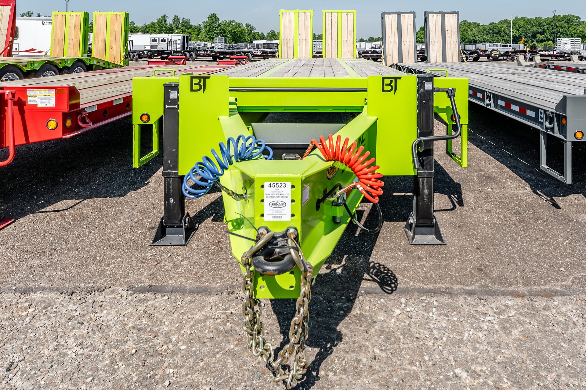 Bright green heavy-duty trailer hitch with blue and red coiled hoses, and hanging chains in the foreground of a truck yard with flatbeds in the background.