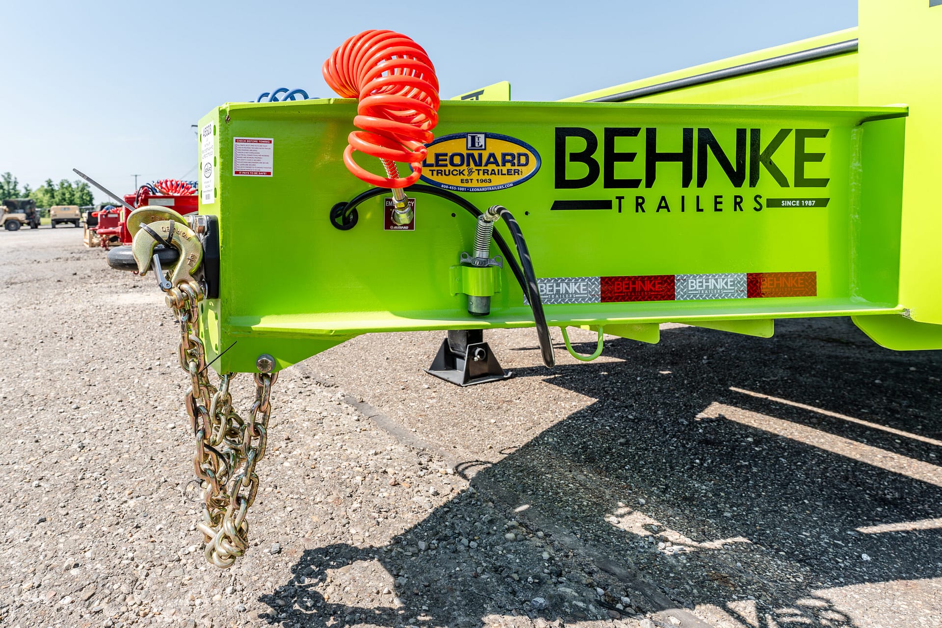 Bright green trailer in a gravel lot with a red coiled air hose and heavy chains at the hitch, Behnke Trailers branding visible.