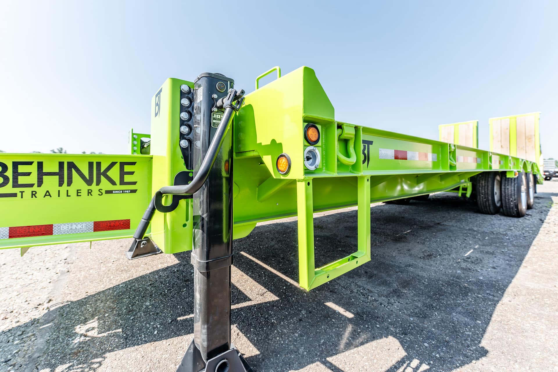 Bright green flatbed trailer with a hydraulic jack and support leg on a gravel lot under a clear blue sky (Behnke Trailers)