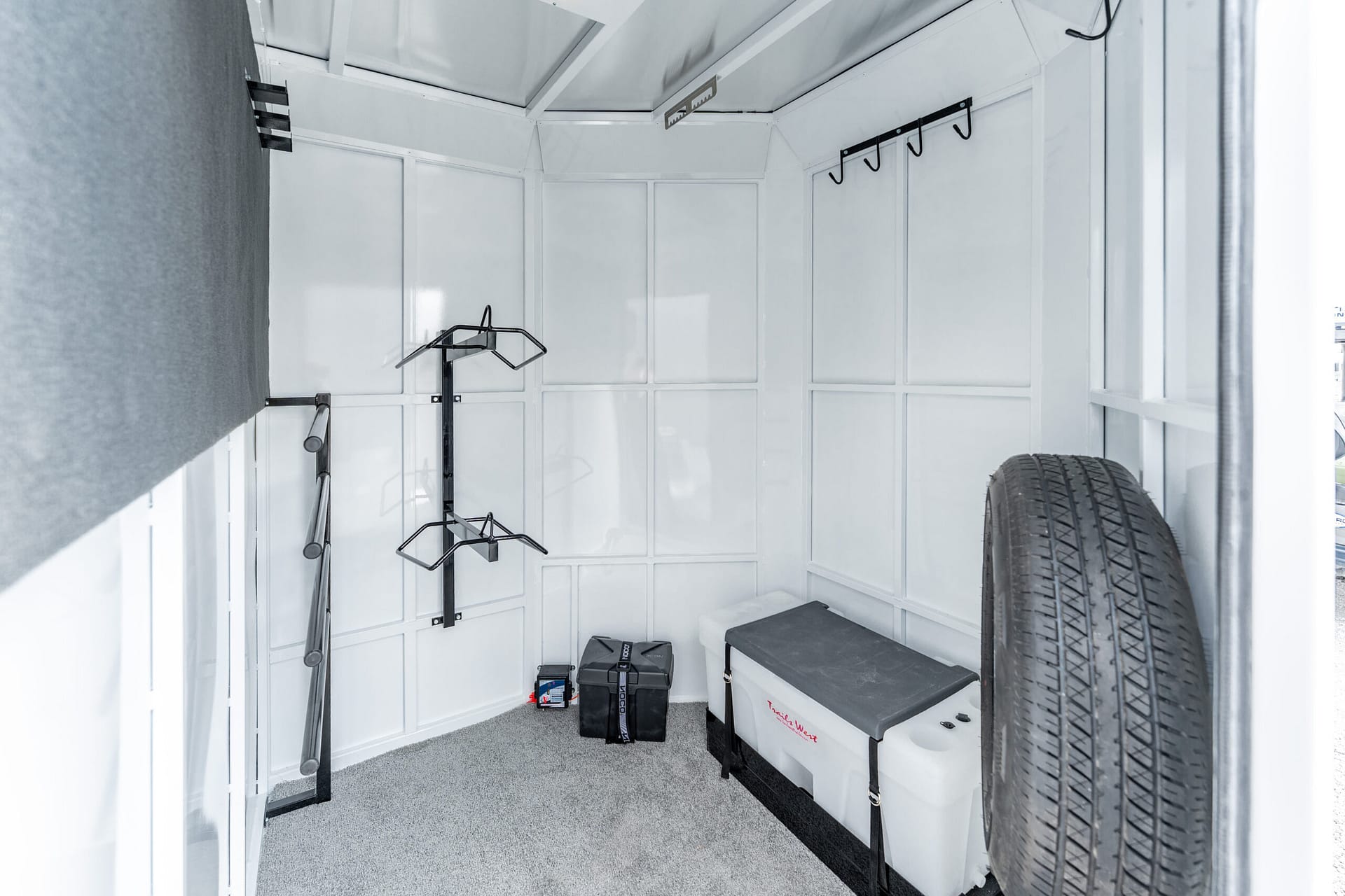Interior of a small white storage trailer with hooks, shelves, a spare tire, and tool boxes on the floor.