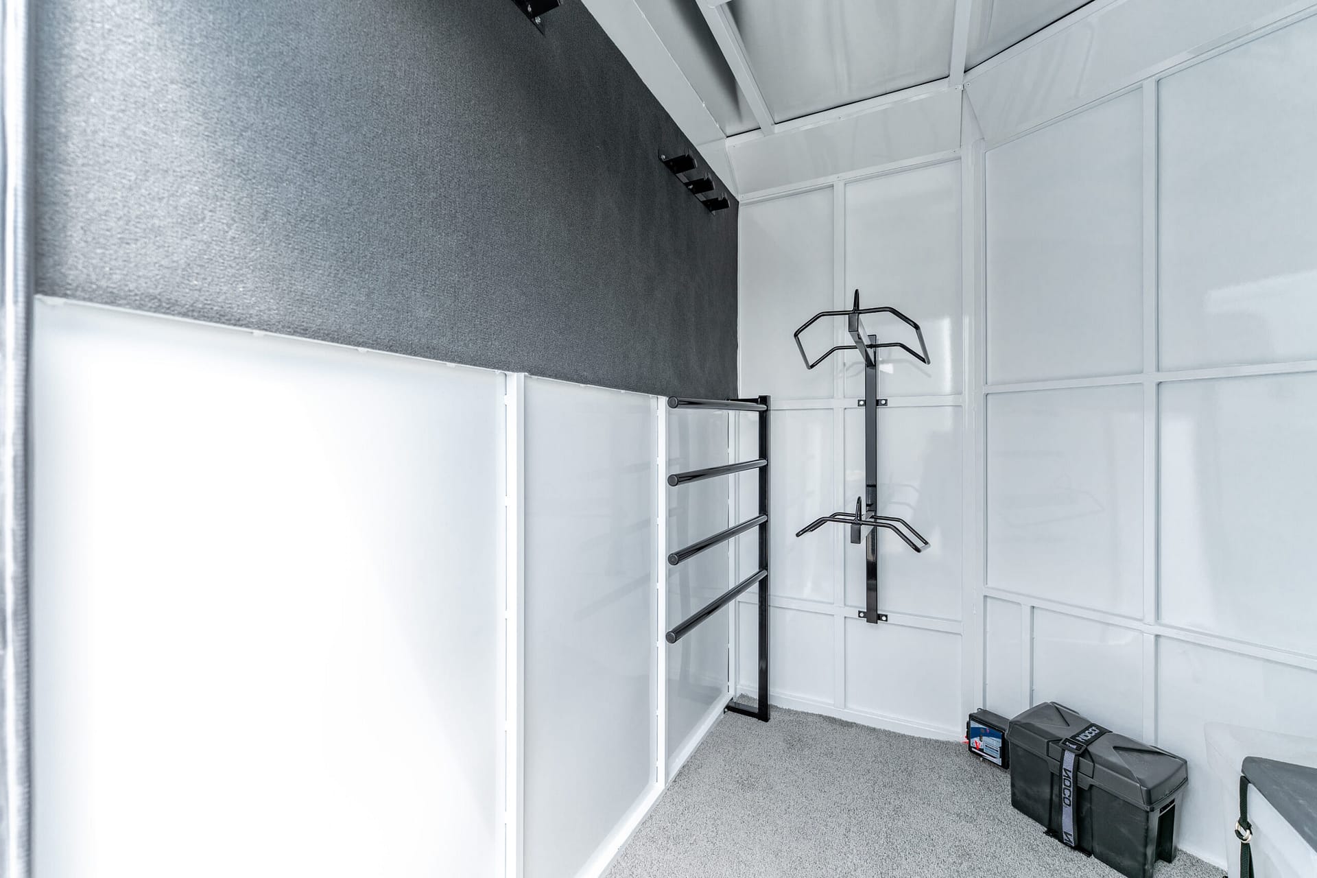 Storage room with white panel walls, metal wall-mounted racks, and a carpeted floor; several black tool boxes sit on the floor to the right.