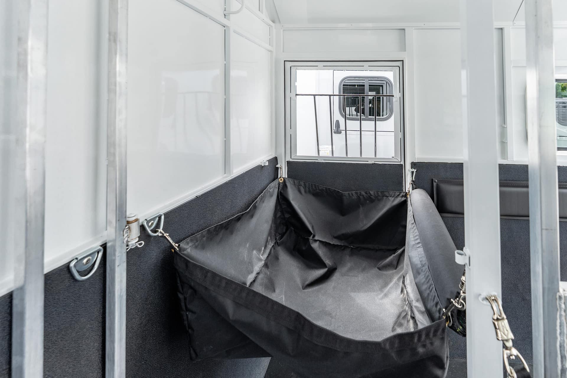 Interior of a white livestock/horse trailer with black fabric bunk bags along the walls and a windowed rear door at the far end.