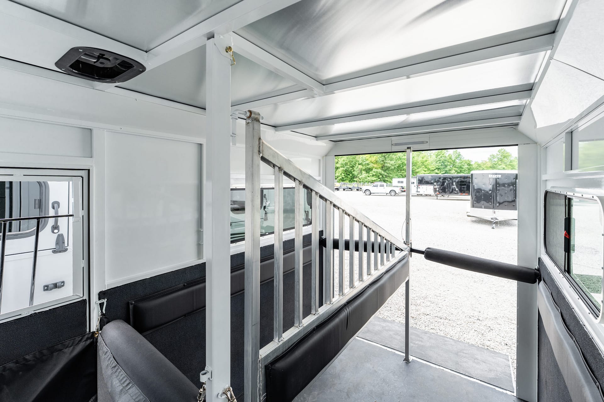 Interior of a metal trailer with a central divider, gray walls, and black padded benches; daylight shows a parking lot outside with other trailers visible.