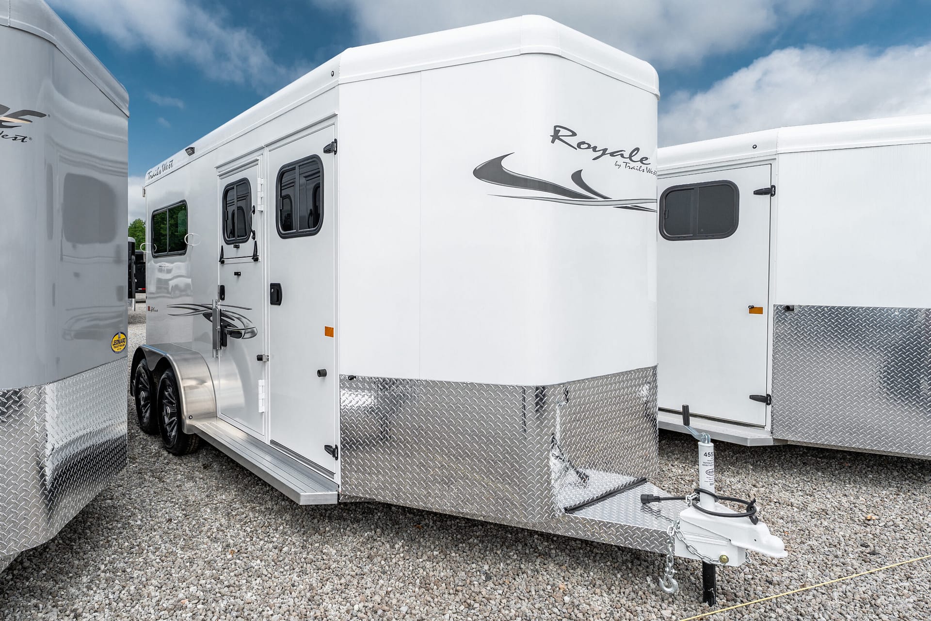 White enclosed trailer with diamond-plate trim and hitch on gravel under a blue sky (side view).