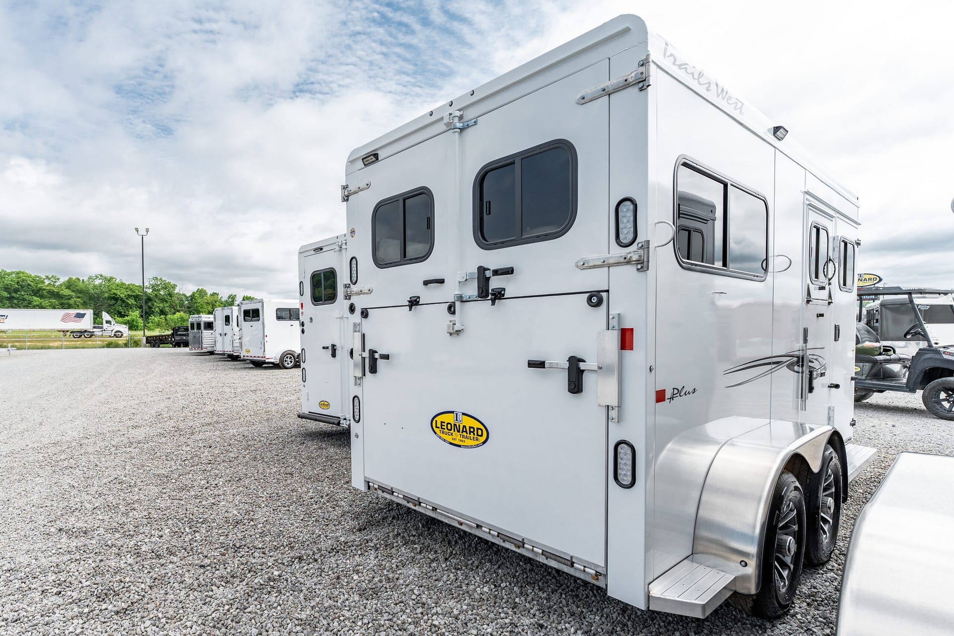 Rear of a white RV trailer with doors, windows, and a Leonard logo visible, parked on a gravel lot with other trailers in the background under a cloudy sky.
