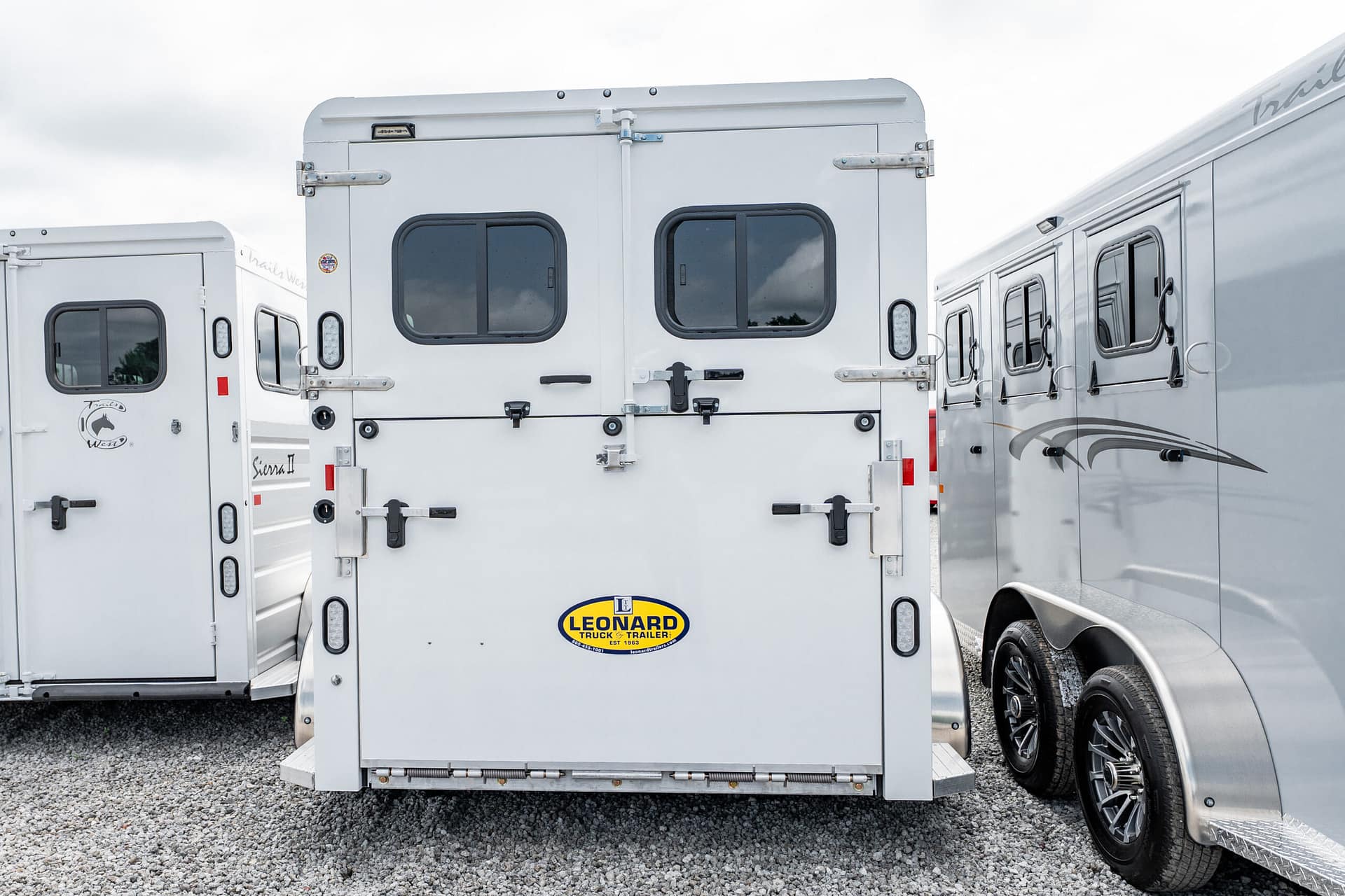 Rear view of a white horse trailer with two back doors and a Leonard Truck & Trailer logo on the bumper, parked among other trailers on a gravel lot.