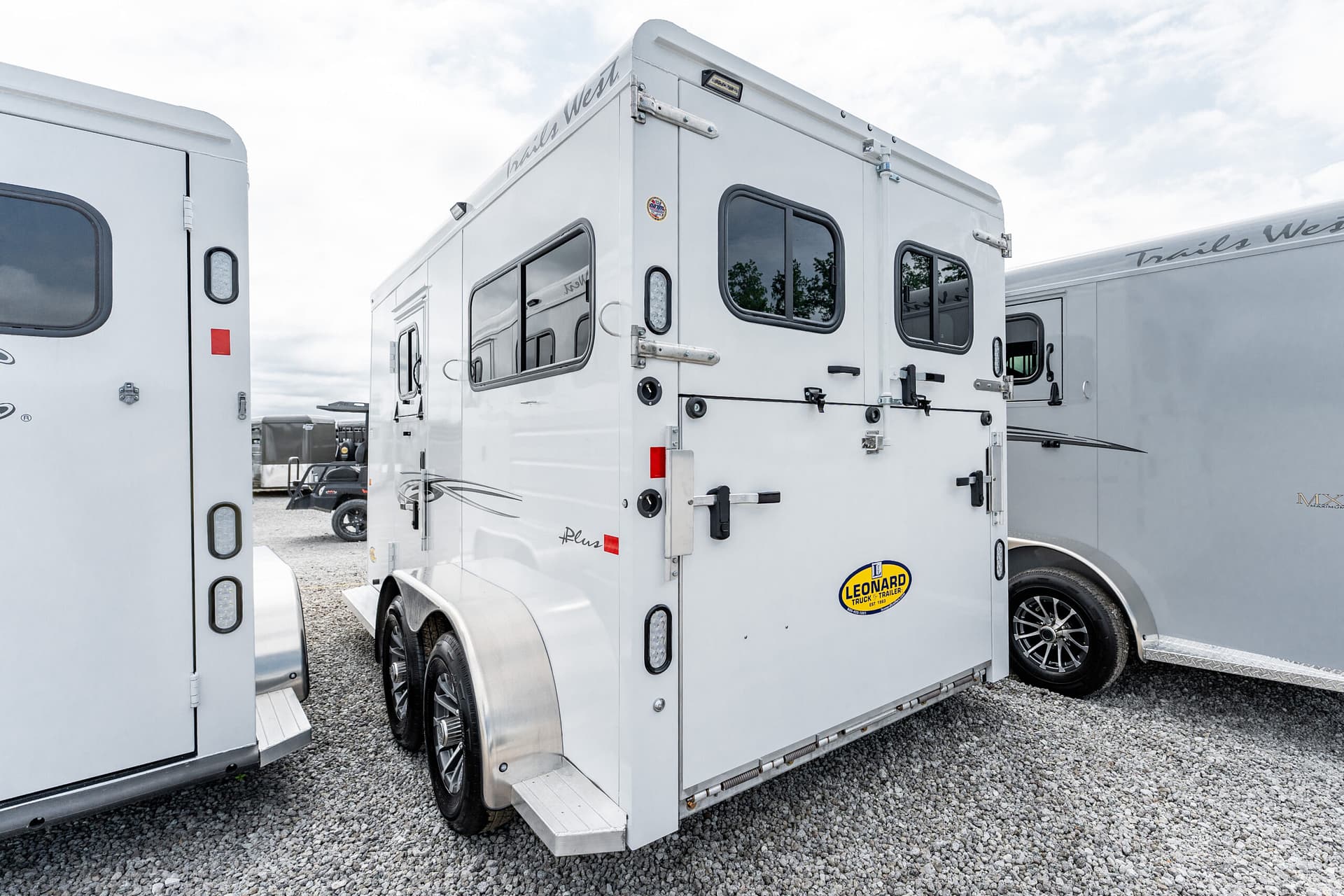 White enclosed trailer with large windows and rear ramp doors parked on a gravel lot; Trail West branding visible on the side and back.