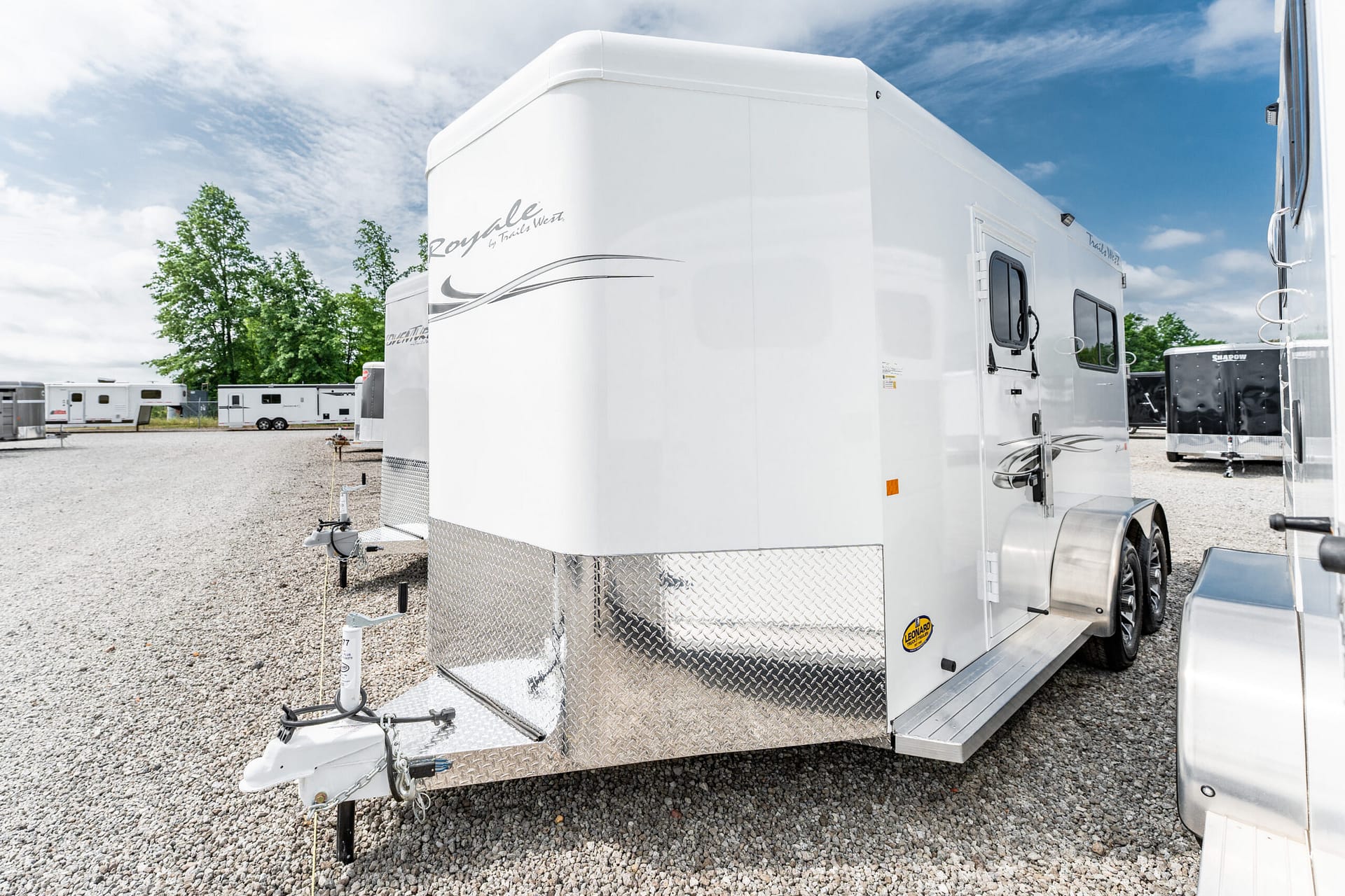 White travel trailer with diamond-plate front parked on a gravel lot, other trailers visible in the background under a blue sky.