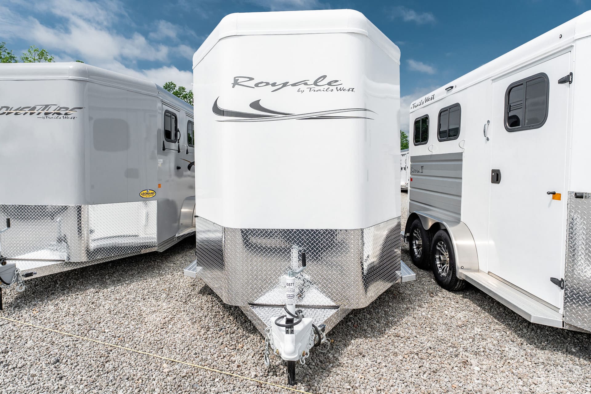 White horse trailer with diamond-plate front, parked between other trailers under a blue sky.