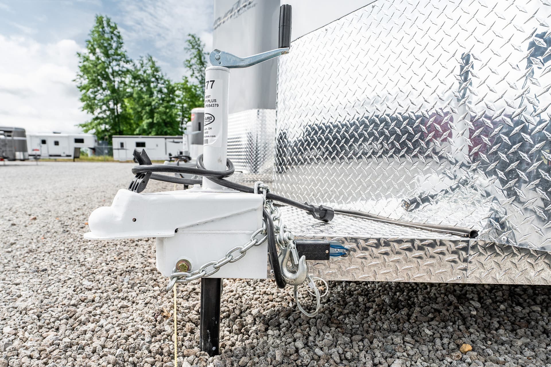 Close-up of a trailer hitch on a diamond-plate trailer, with a white jack, safety chains, and cables on a gravel lot.