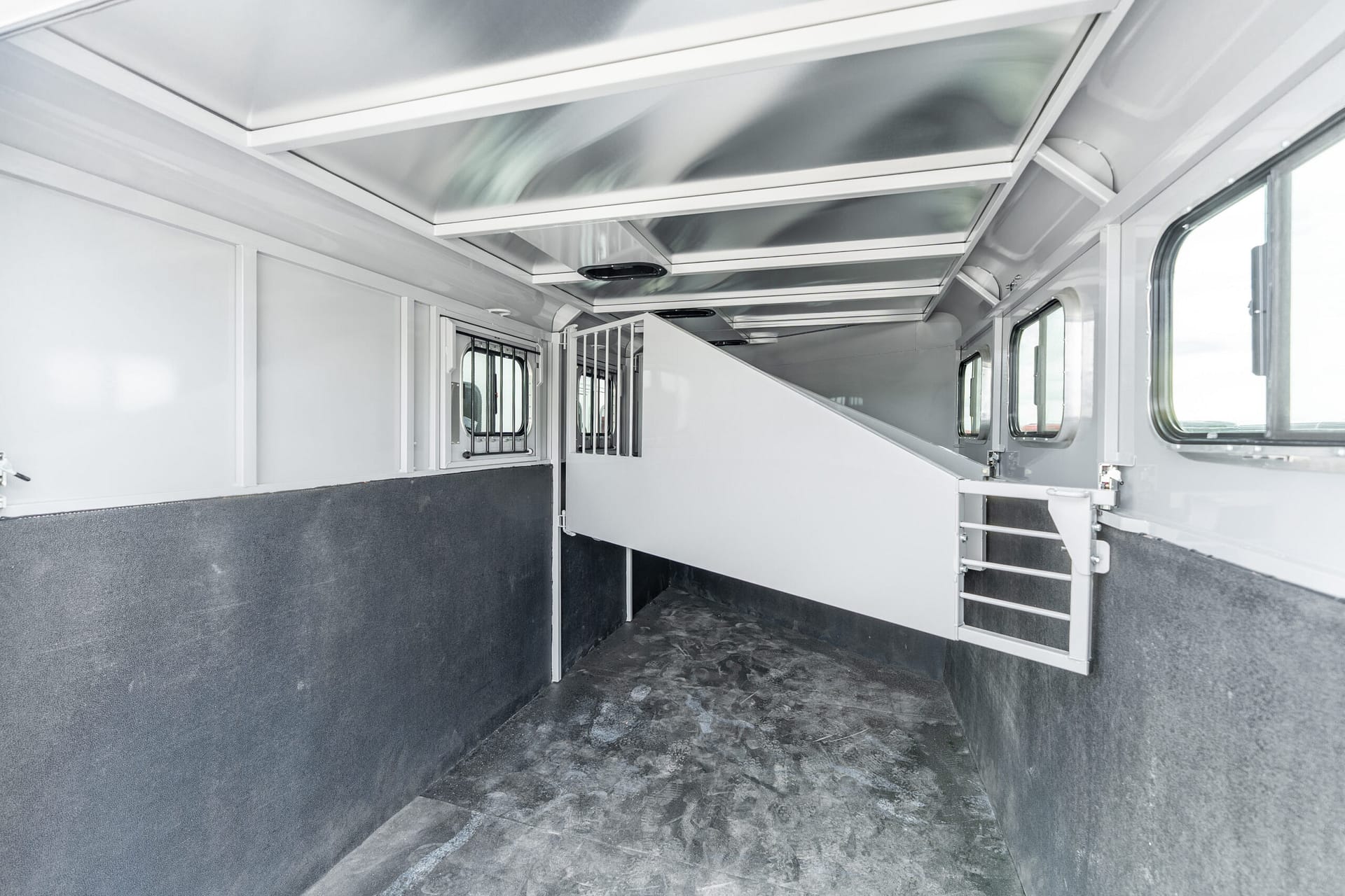 Interior of a light gray horse trailer with divider stalls, small barred windows, and a textured rubber floor.