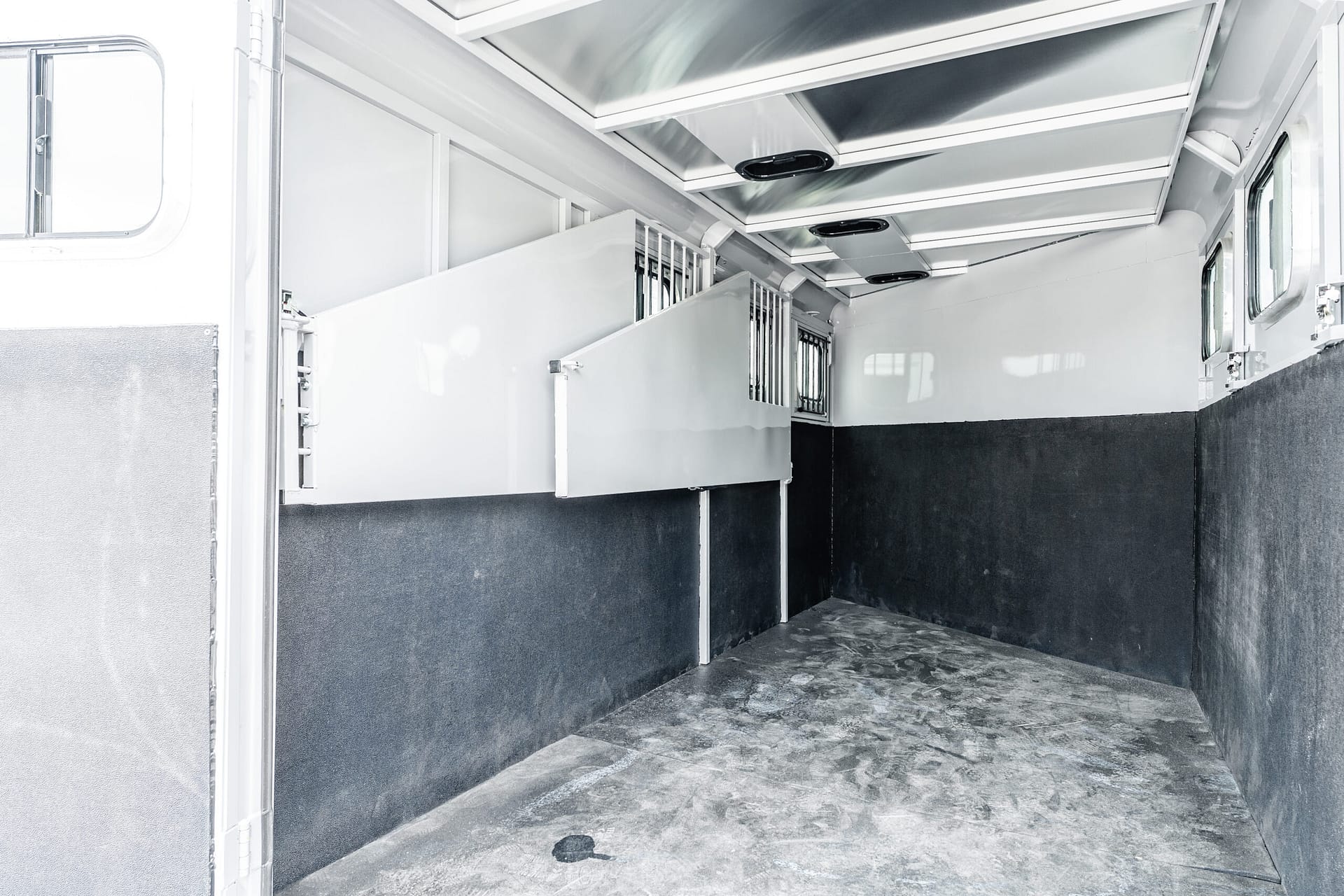 Interior of an empty horse trailer with metal divider bars and two stalls, white upper walls, black lower panels, and a rubber floor.