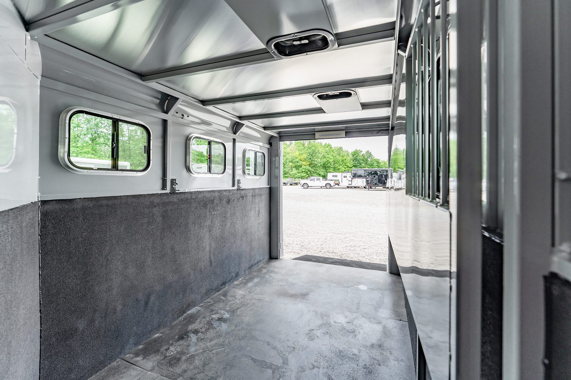 Interior of a silver trailer with side windows and a view of a parking lot outside with trees and vehicles outside the doorway reading light gray walls and a dark lower panel.