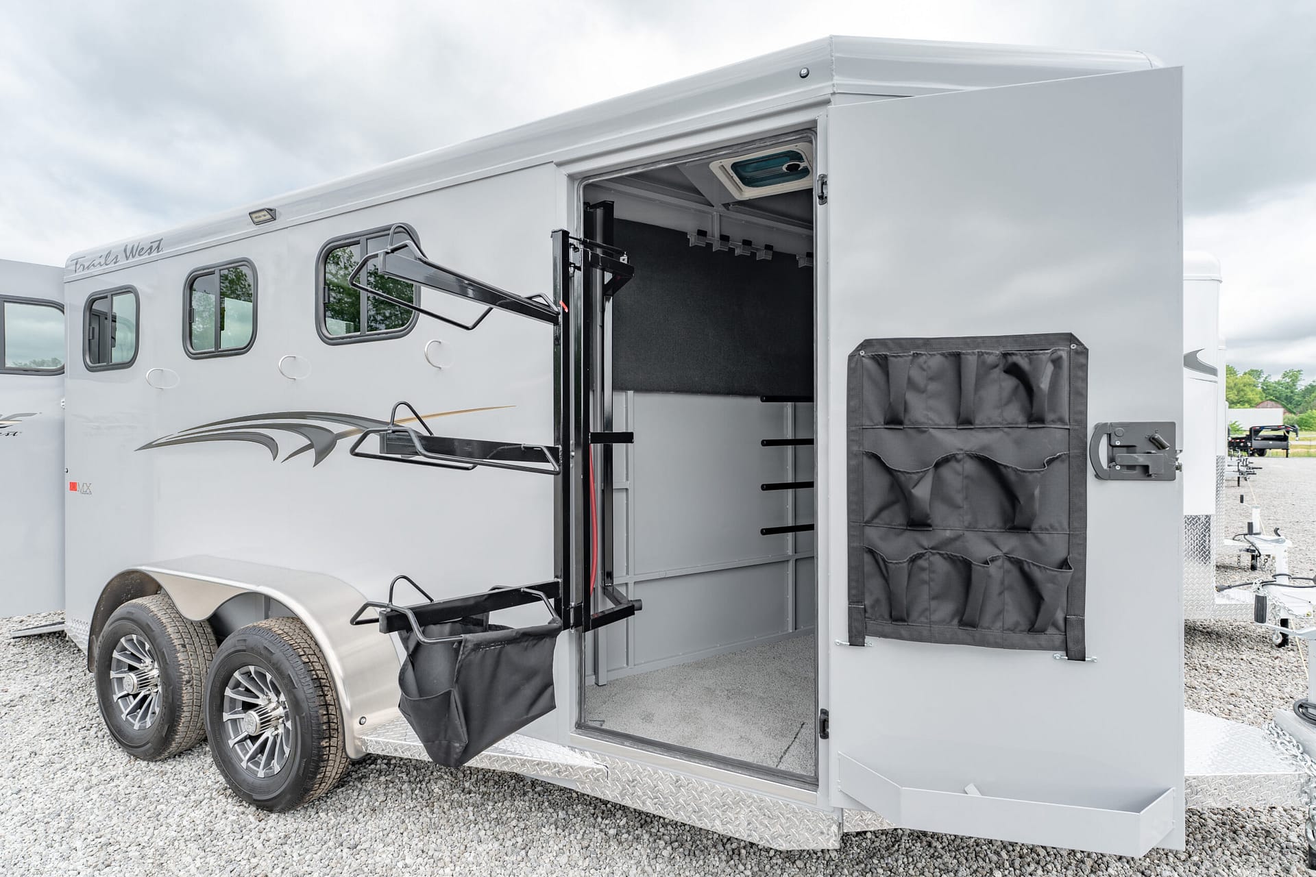 Open side of a silver horse trailer with interior storage visible and external hanging pouches on the door, parked on gravel under cloudy sky.