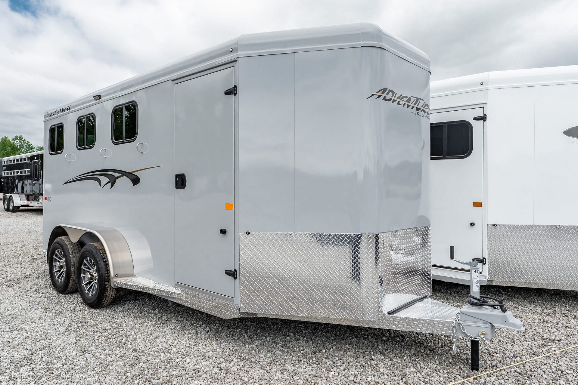 Silver enclosed trailer with rounded front, two axles and diamond-plate trim parked on a gravel lot.