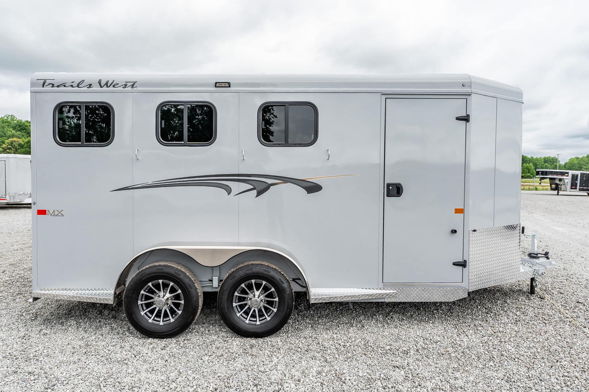 Grey Trails West horse trailer seen from the side with three rounded windows, decorative swoosh graphics, and tandem wheels on a gravel lot.