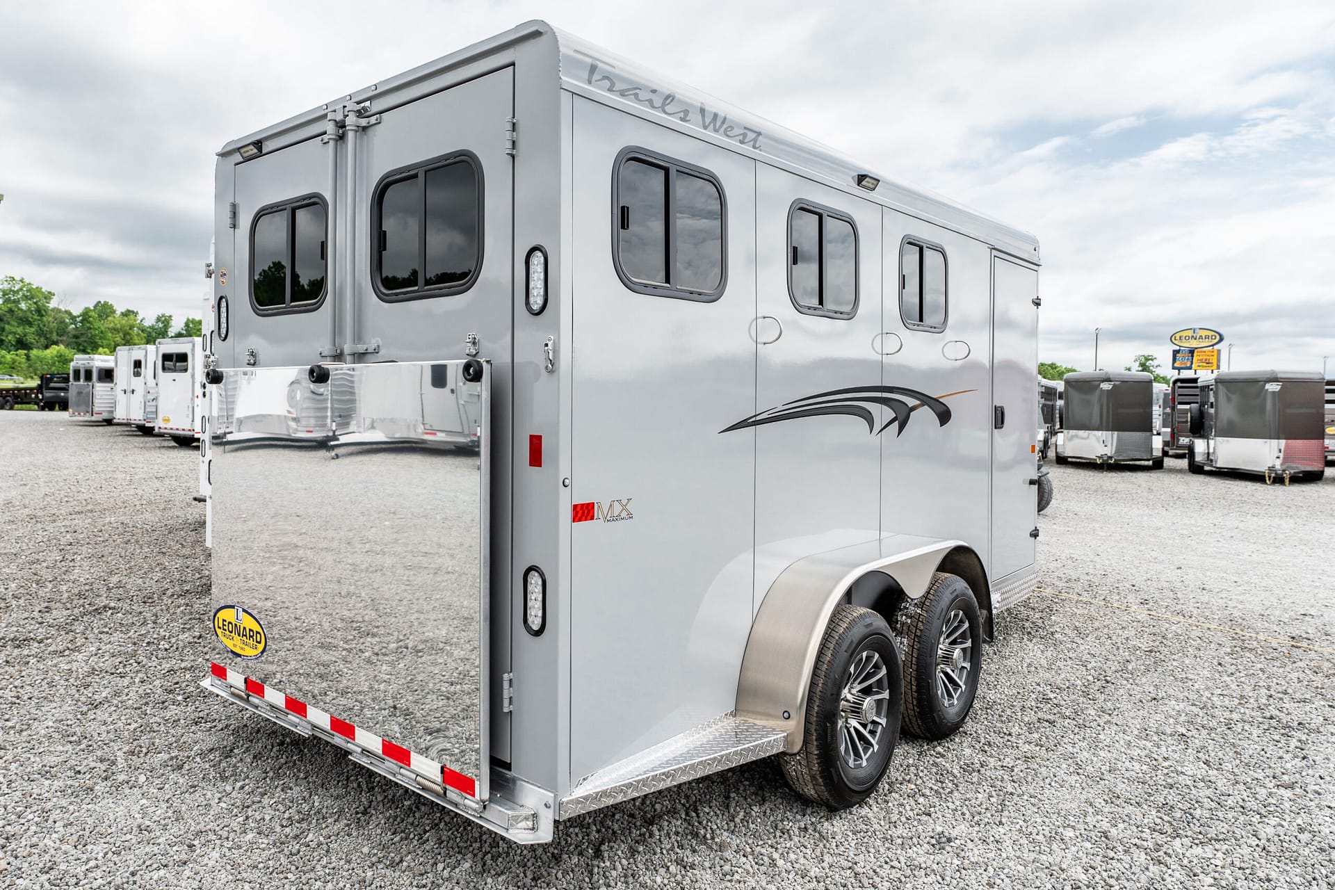 Rear and side view of a large silver trailer with windows, parked on a gravel lot at a trailer dealer. The Leonard sign is visible in the background.
