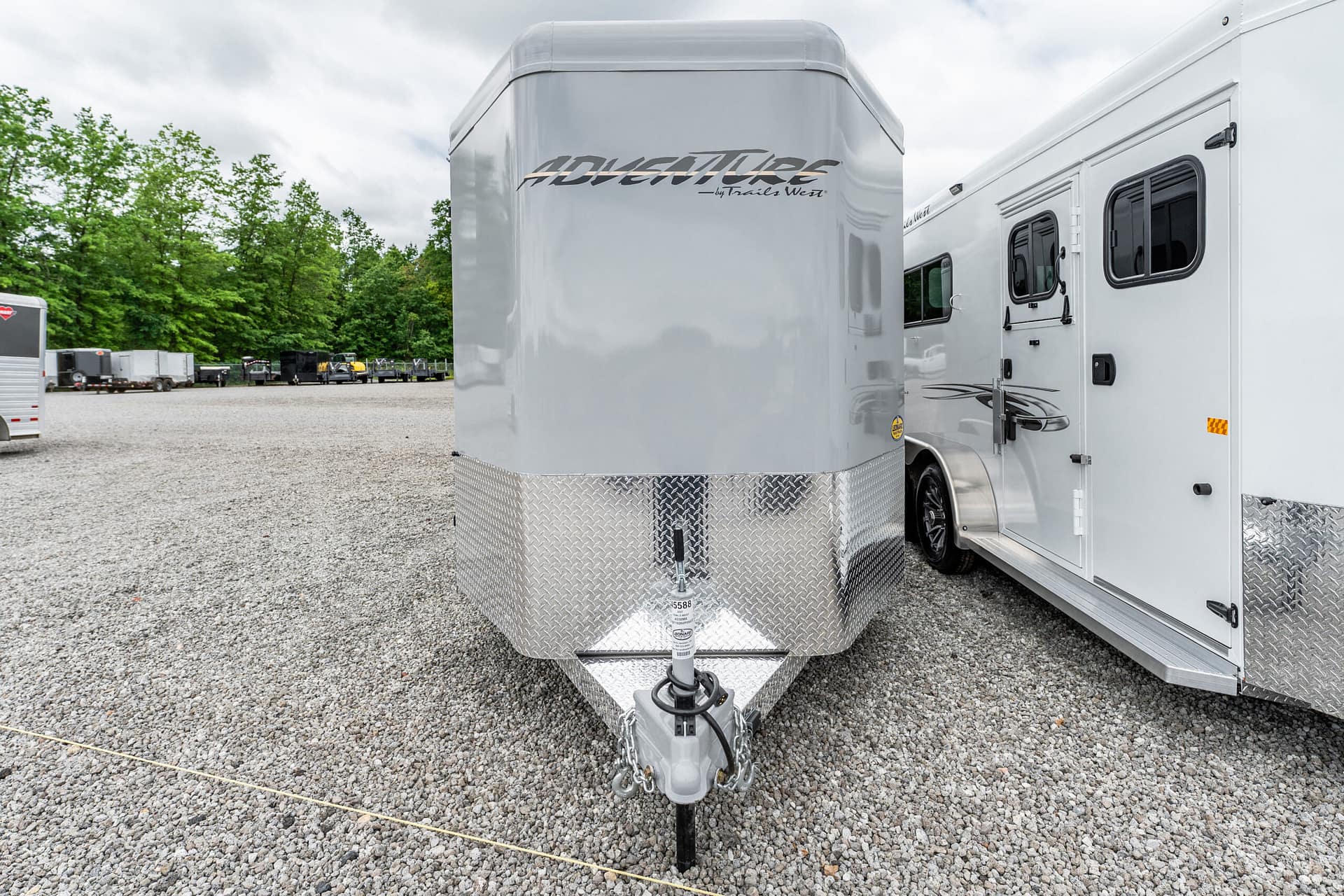 Silver enclosed trailer with diamond-plate front, hitch, and towing hitch visible in a gravel lot with trees in the background.