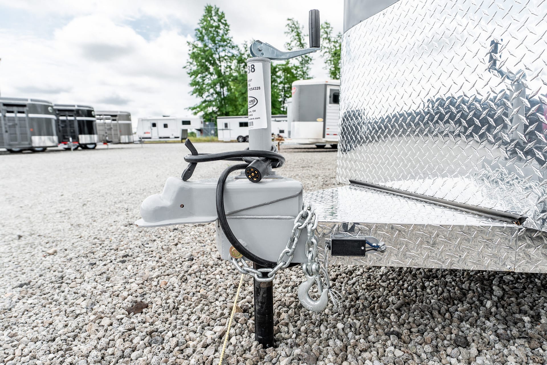 Close-up of a trailer hitch assembly with a safety chain and electrical plug on a diamond-plate trailer, set on a gravel lot with other trailers in the background.