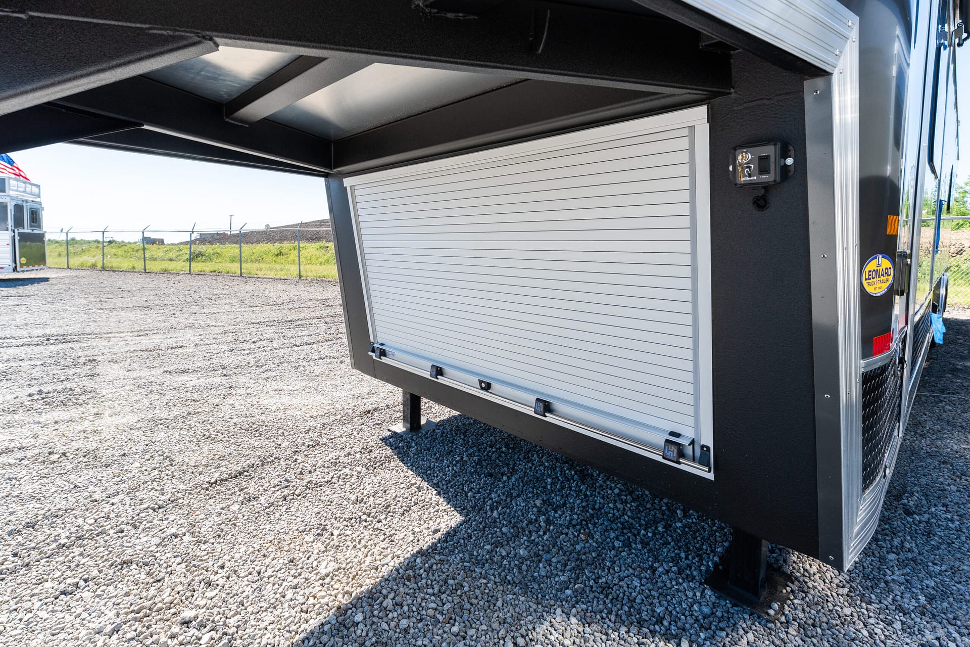 Black trailer exterior with a silver ribbed roll-up storage door, parked on a gravel lot; a chain-link fence and distant American flag are visible in the background.