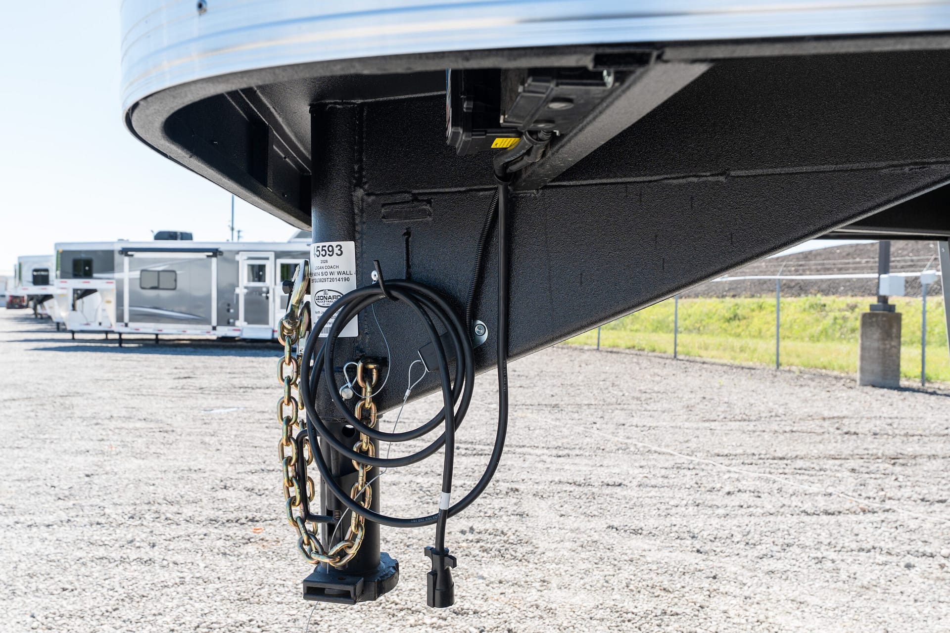 Close-up of a trailer hitch with connected cables and safety chains hanging from a black metal frame, in a lot with other trailers in the background.