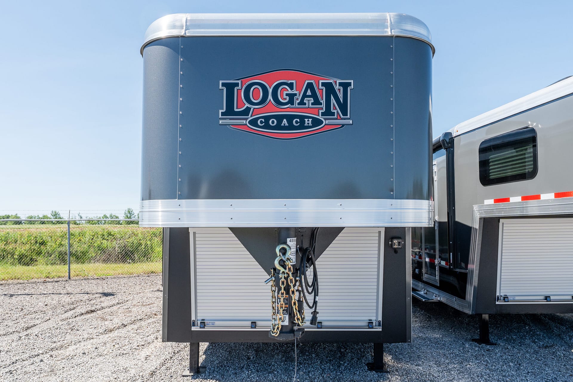 Front panel of a blue Logan Coach trailer with safety chains hanging, silver trim, and part of another trailer visible on the right.