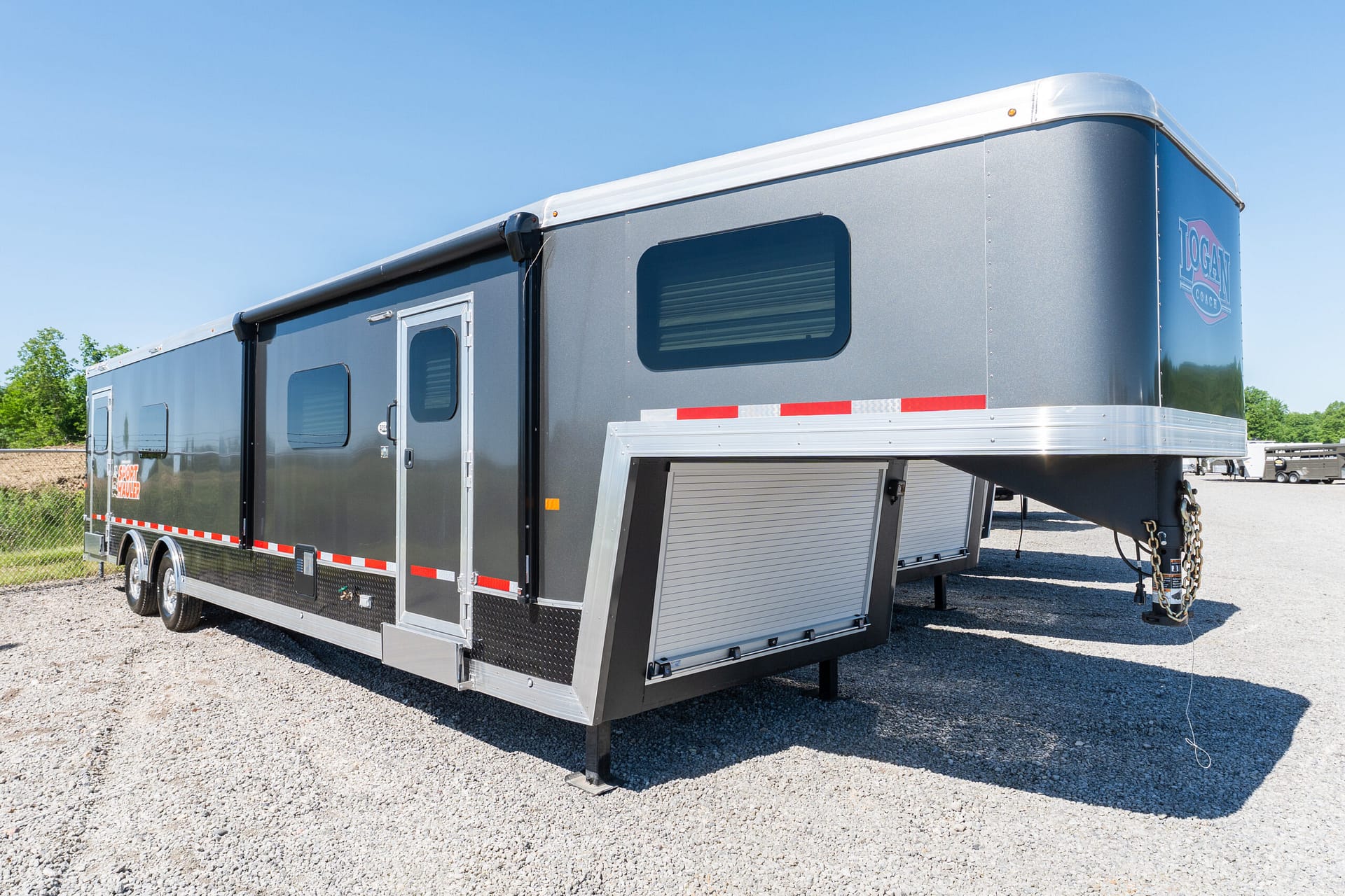 Gray gooseneck horse trailer with side window and storage compartments parked on a gravel lot beneath a clear blue sky, logo visible on the rear side panel