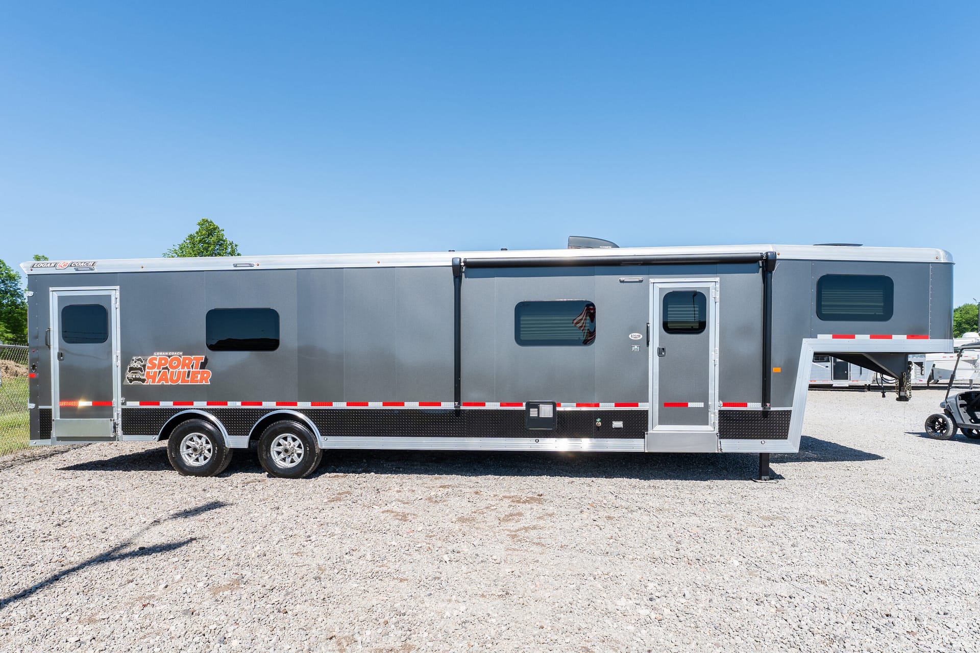 Gray sport-hauler trailer on a gravel lot under a clear blue sky, with 'Sport Hauler' logo on the side.