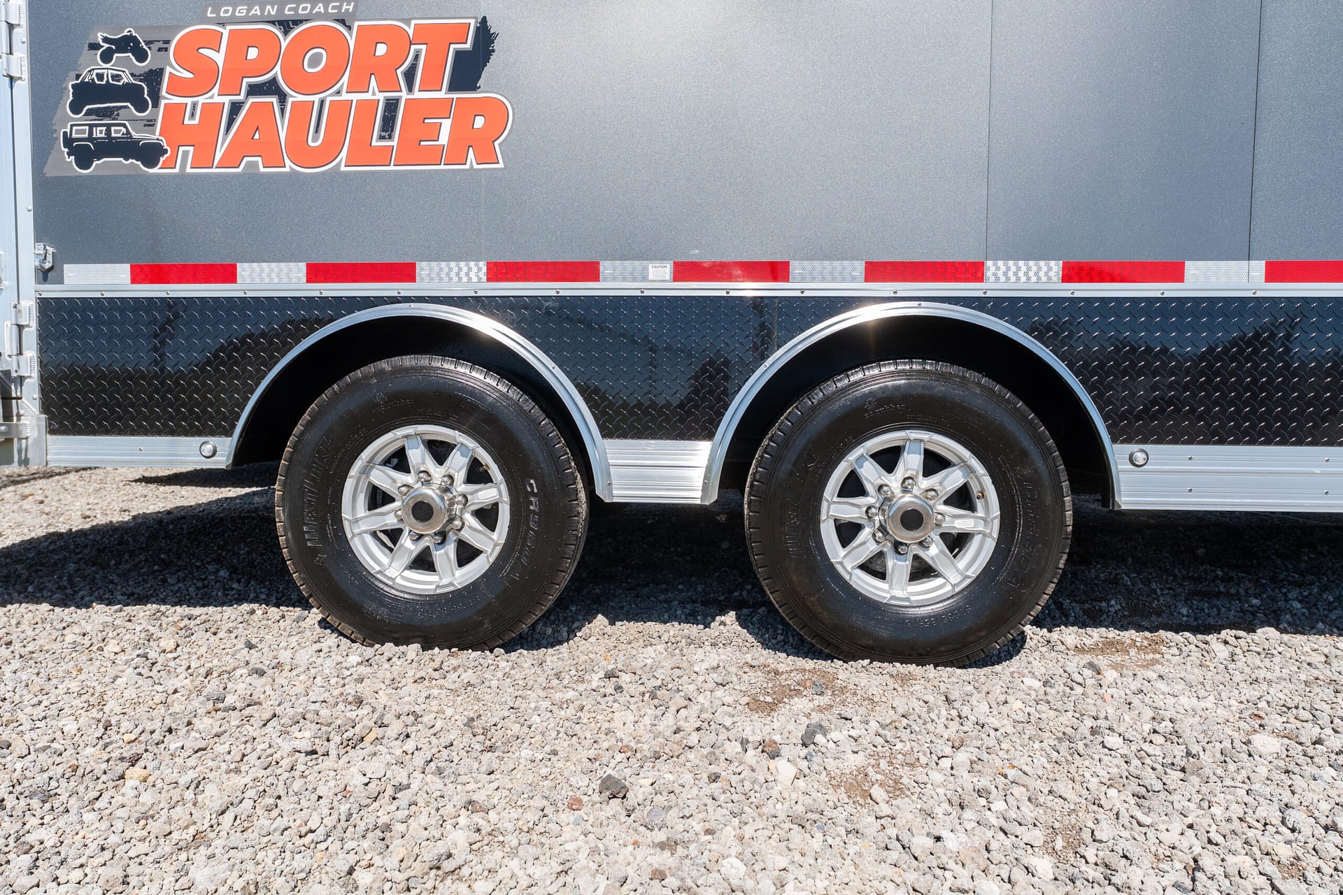 Side of a gray Sport Hauler trailer with large orange logo, diamond-plate lower panel, and dual wheels on gravel undercarriage.