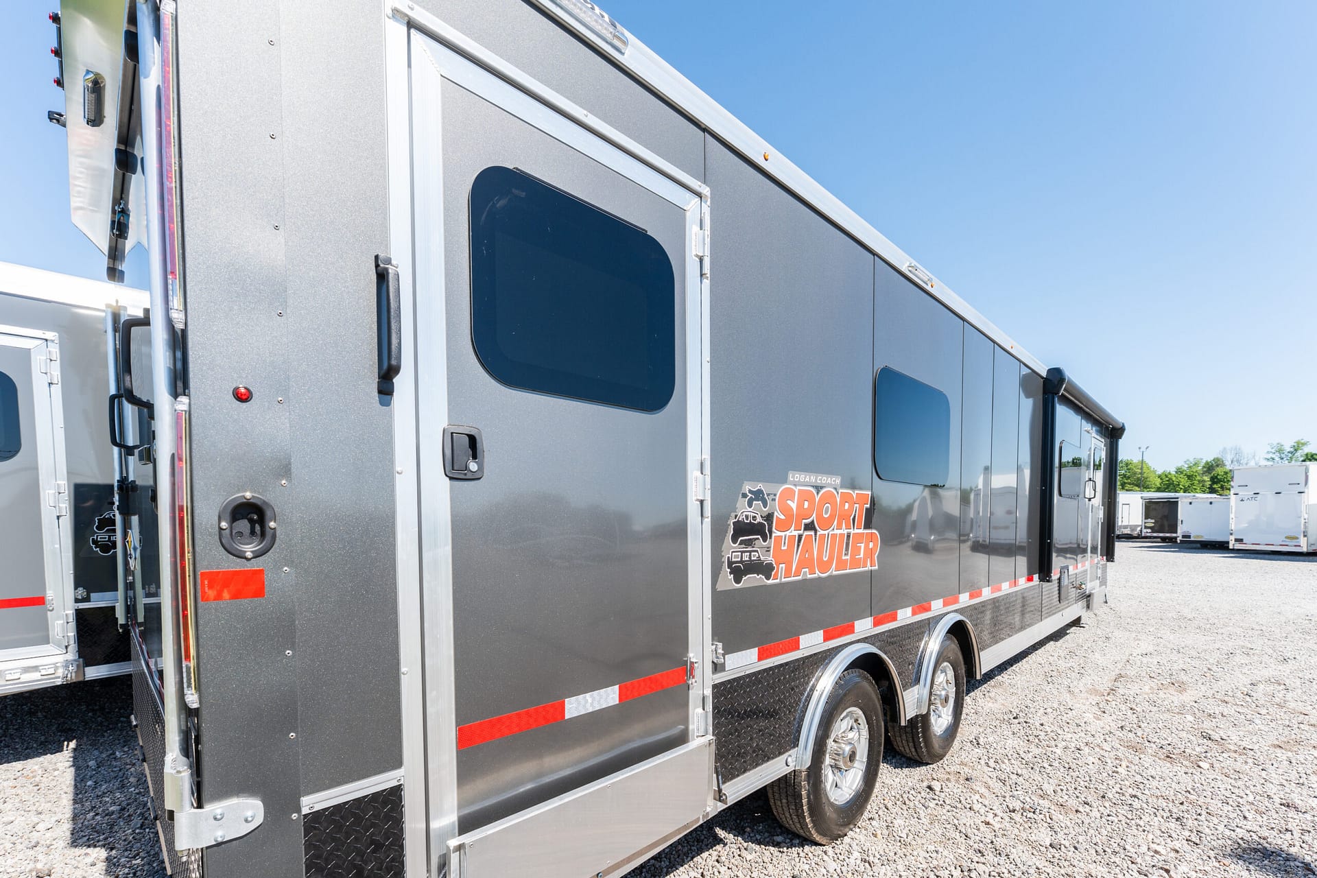 Gray sport hauler trailer parked on a gravel lot under a clear blue sky, side view showing windows and doors with red reflectors.