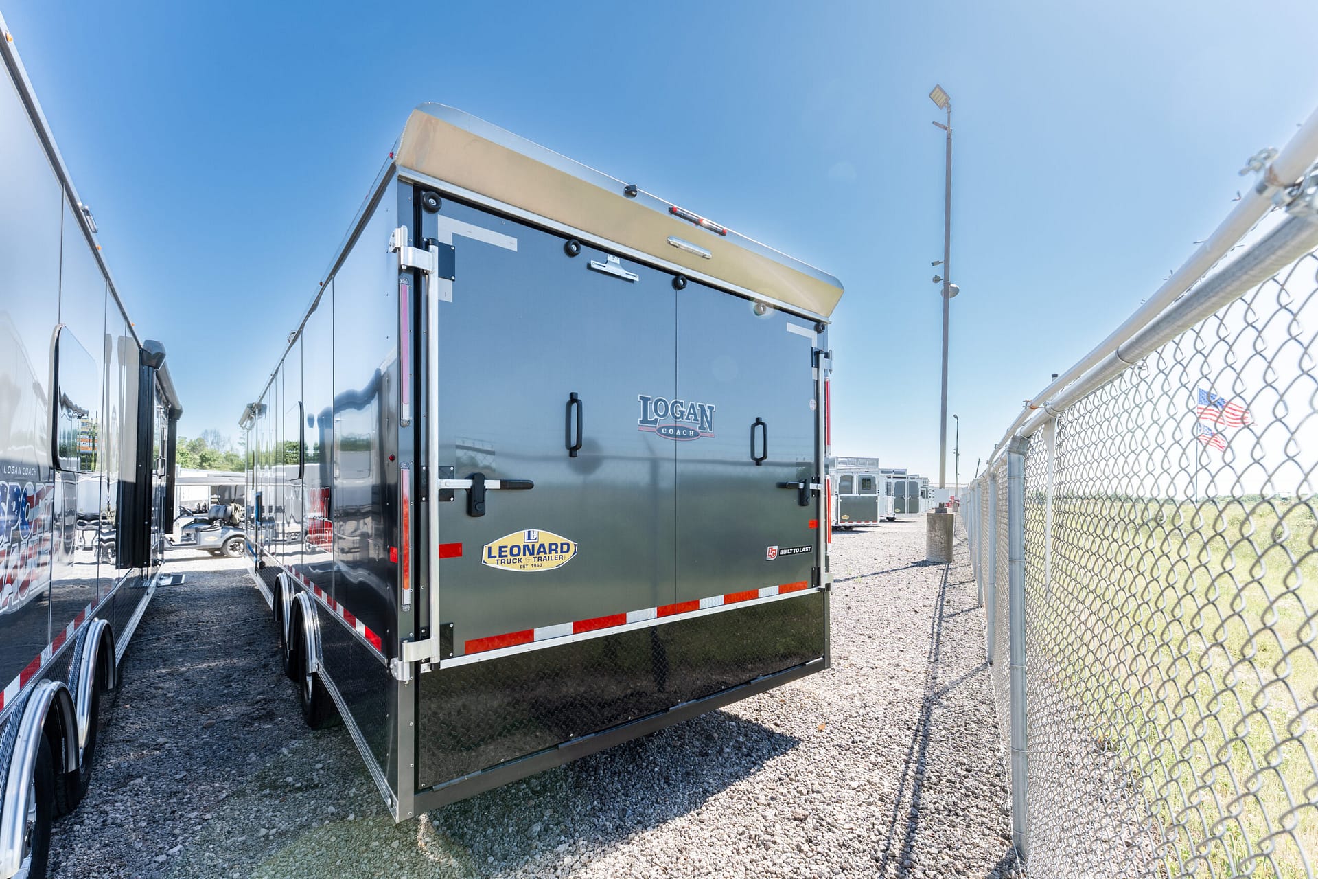 Back of a gray Logan Coach trailer in a row of trailers, chain-link fence on the right, clear blue sky above.