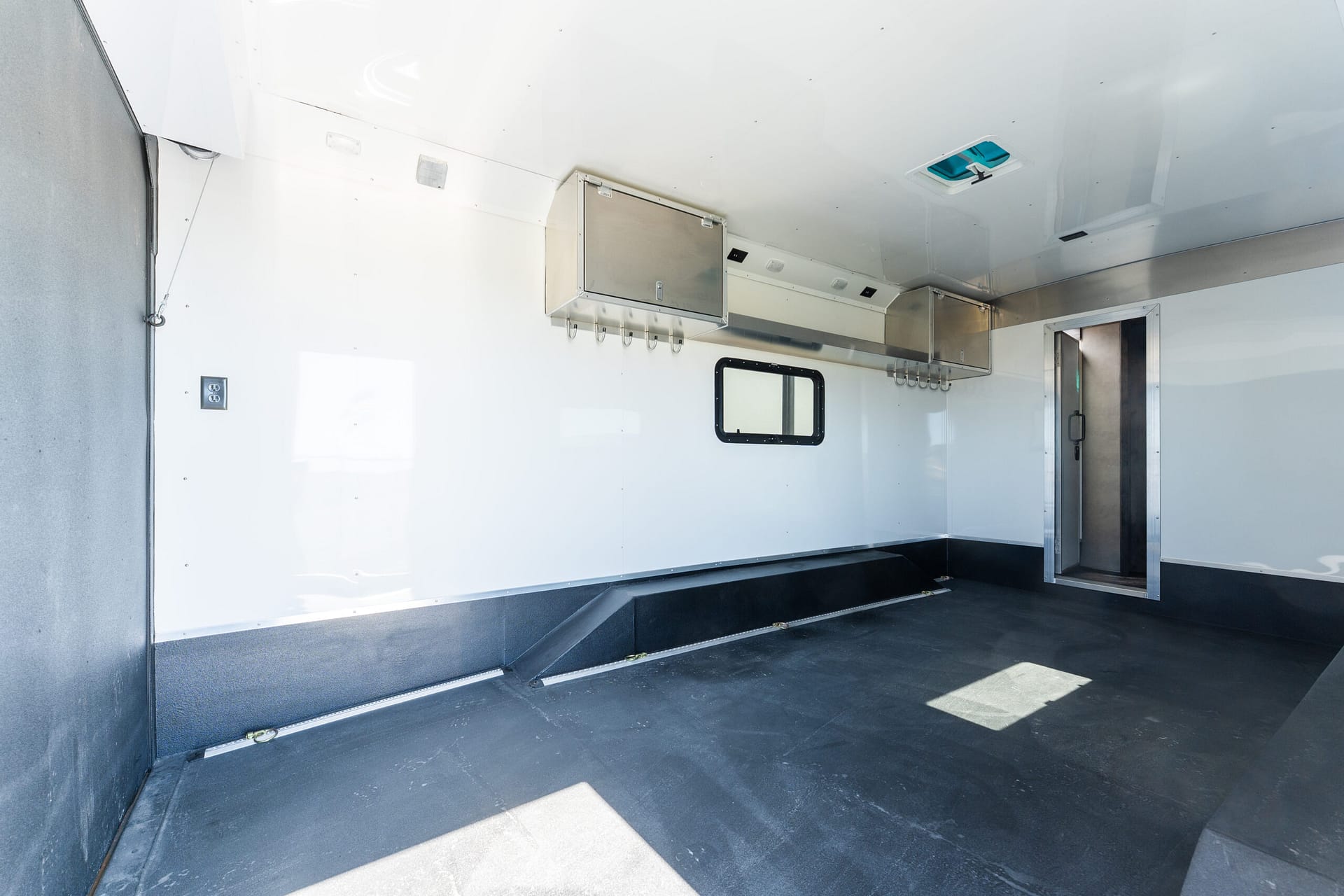 Interior of a white trailer with a hanging metal hatch and row of hooks along the wall, small rectangular window visible on the side wall