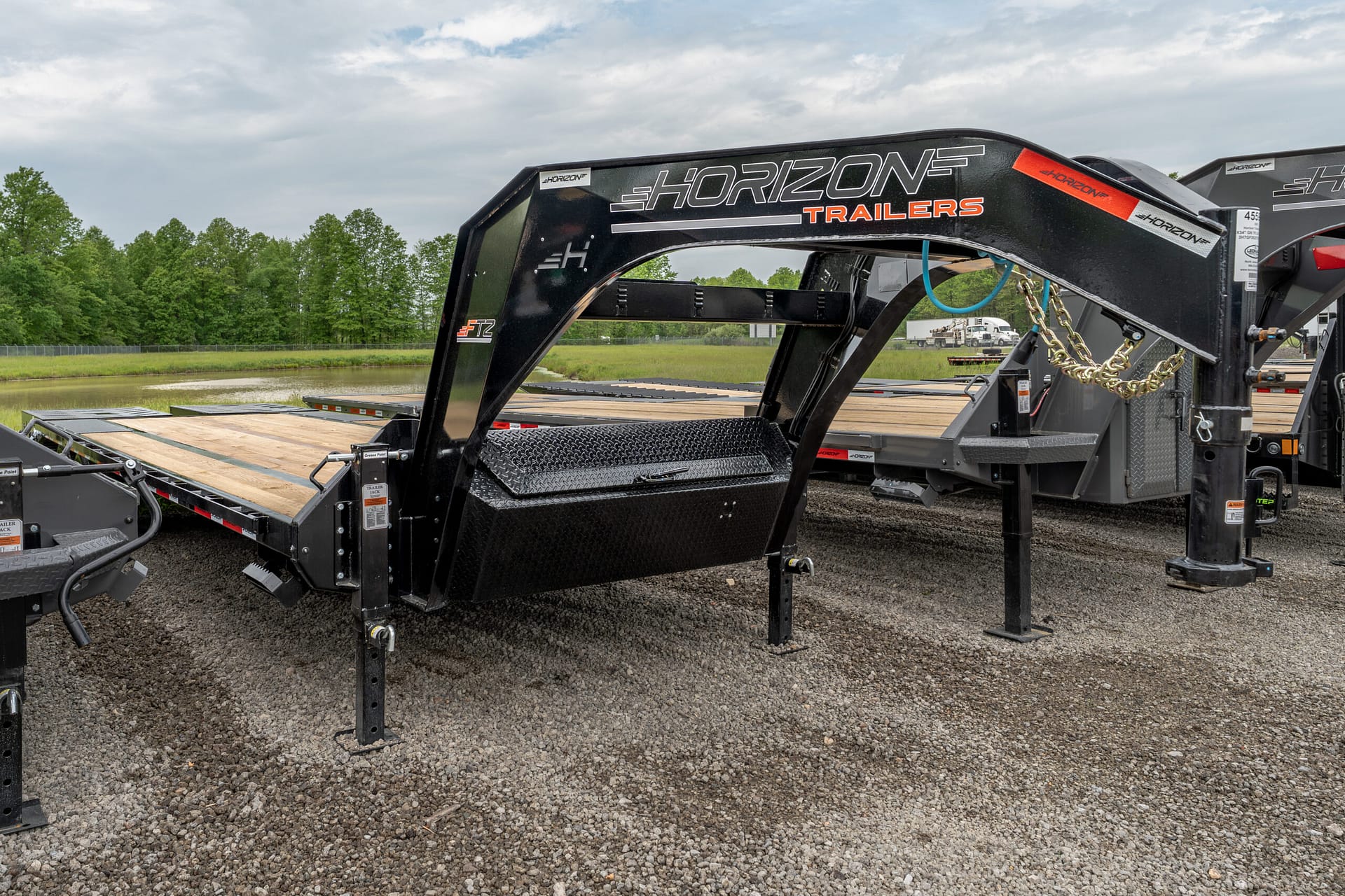 Gooseneck trailer hitch frame branded Horizon Trailers on a gravel lot, with trees and a waterway in the background.