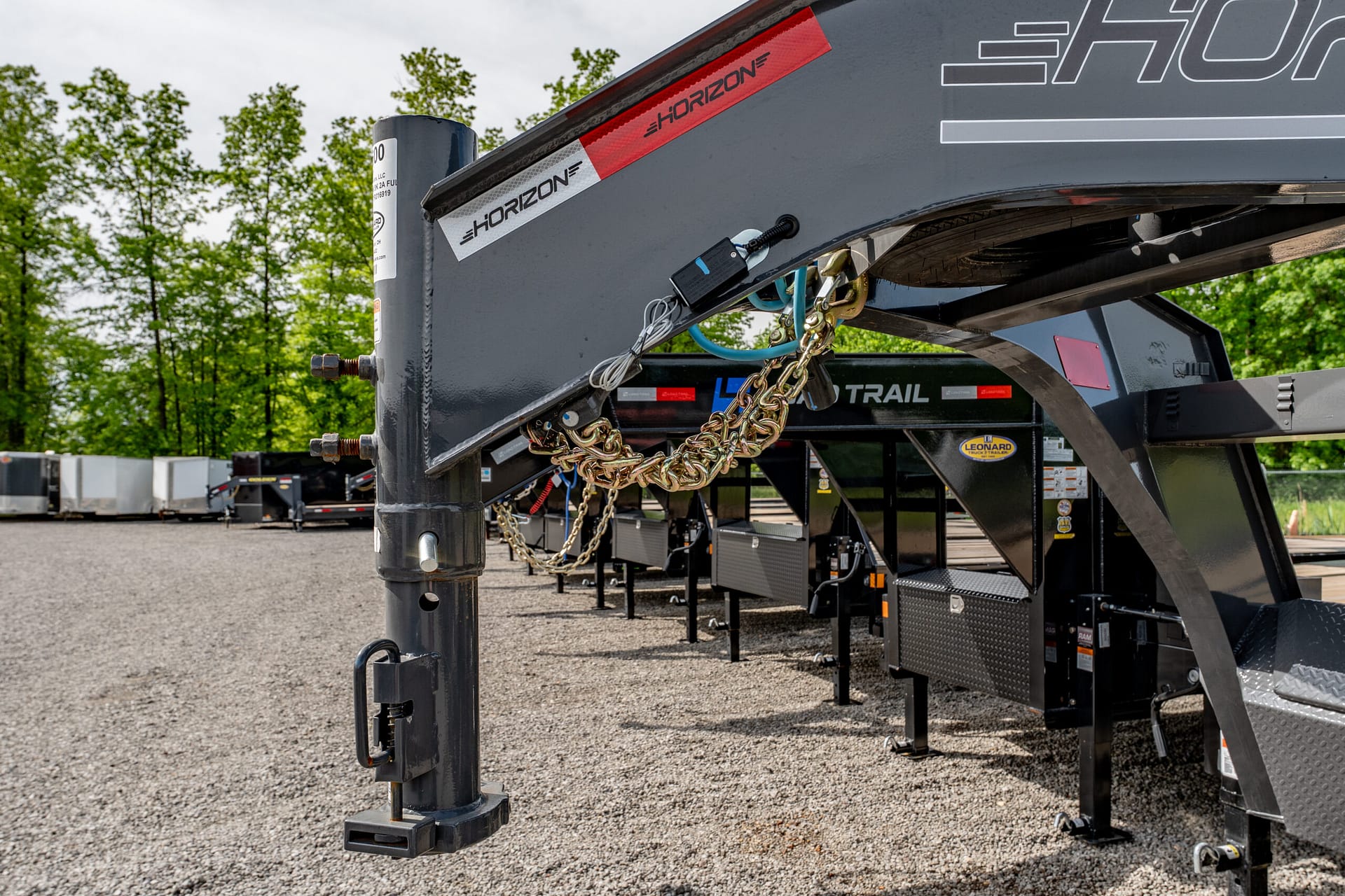 Close-up of a trailer hitch and safety chains on a gray equipment trailer, with a row of other trailers in the background.